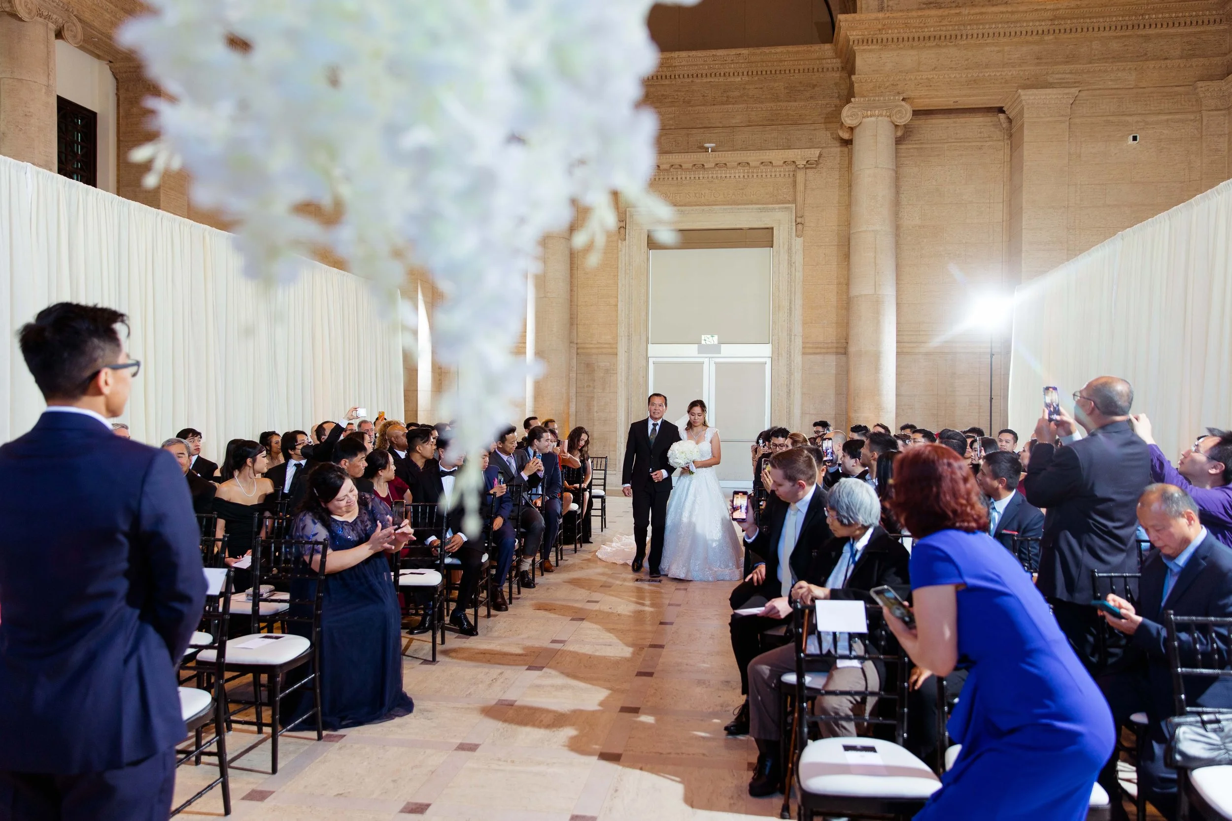 A bride walking down the aisle with her father at a wedding ceremony, with guests seated on both sides capturing the moment.