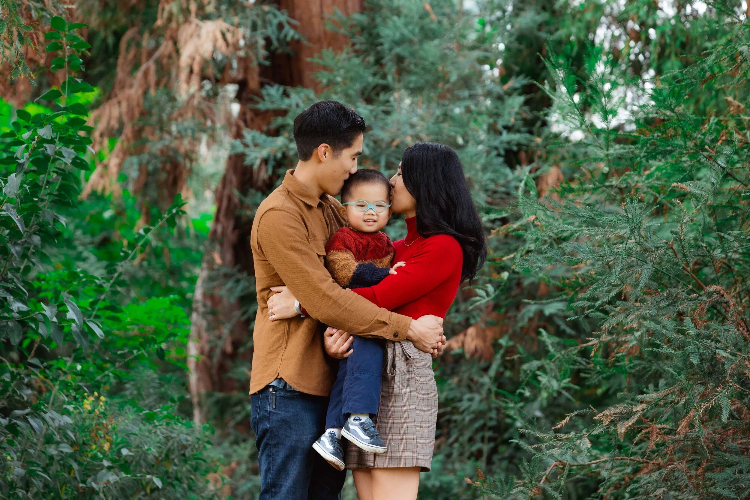 A family of three standing close together in a green outdoor setting, with a man and woman gently kissing a young boy who is smiling and wearing glasses.