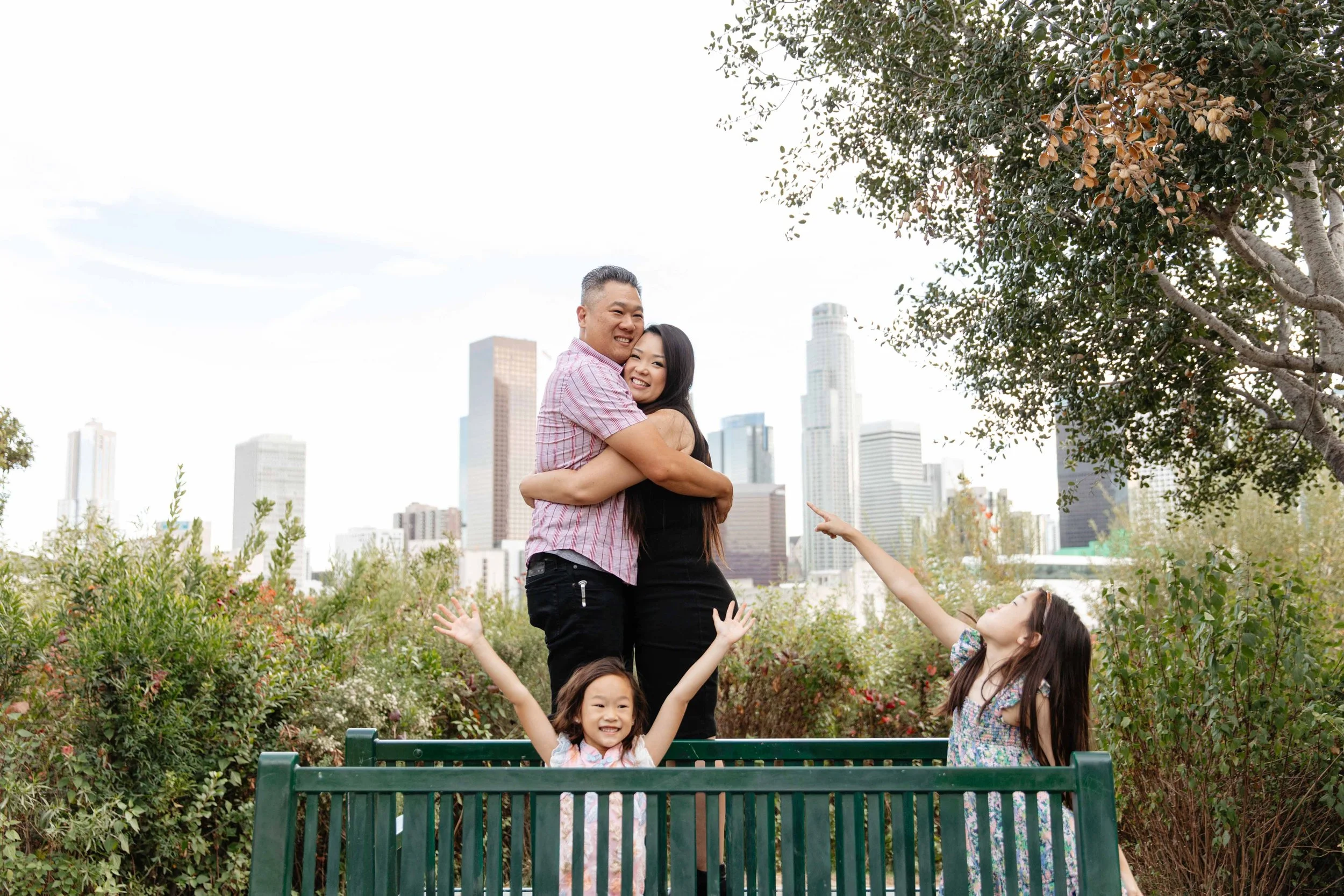 A family of four, consisting of a father, mother, and two young daughters, is enjoying a moment on a park bench in an urban area with skyscrapers in the background. The father and mother are hugging, and the daughters are standing in front with their