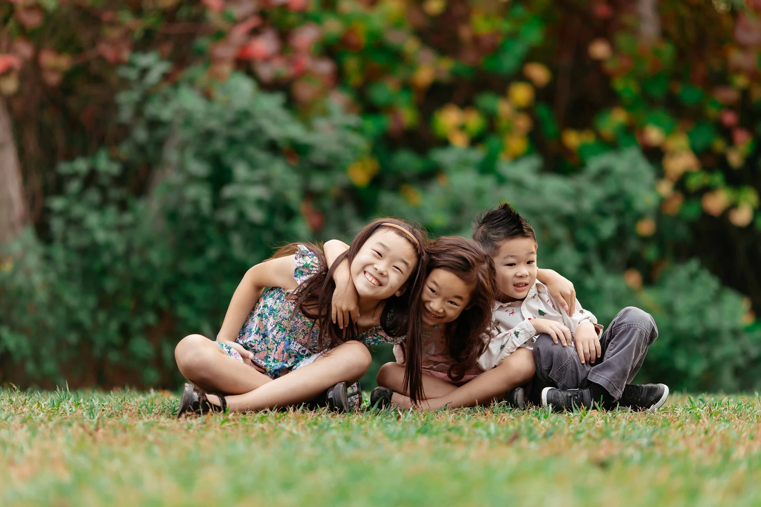 Three children sitting on grass outdoors, smiling and hugging each other, with a blurred green and colorful background.