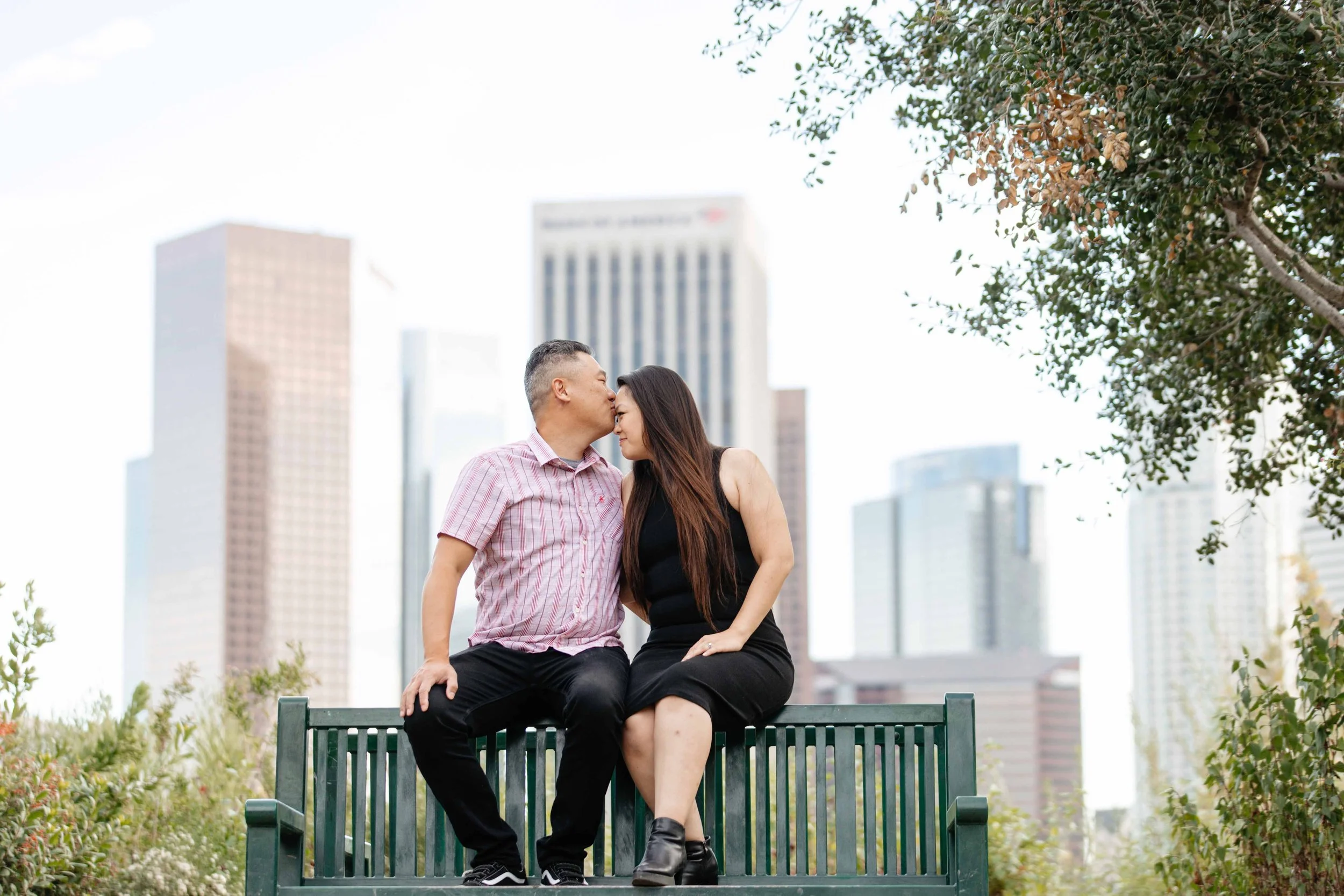 A couple sitting on a park bench in an urban area, sharing a tender moment with the man kissing the woman's forehead, with tall city buildings in the background and trees on the sides.