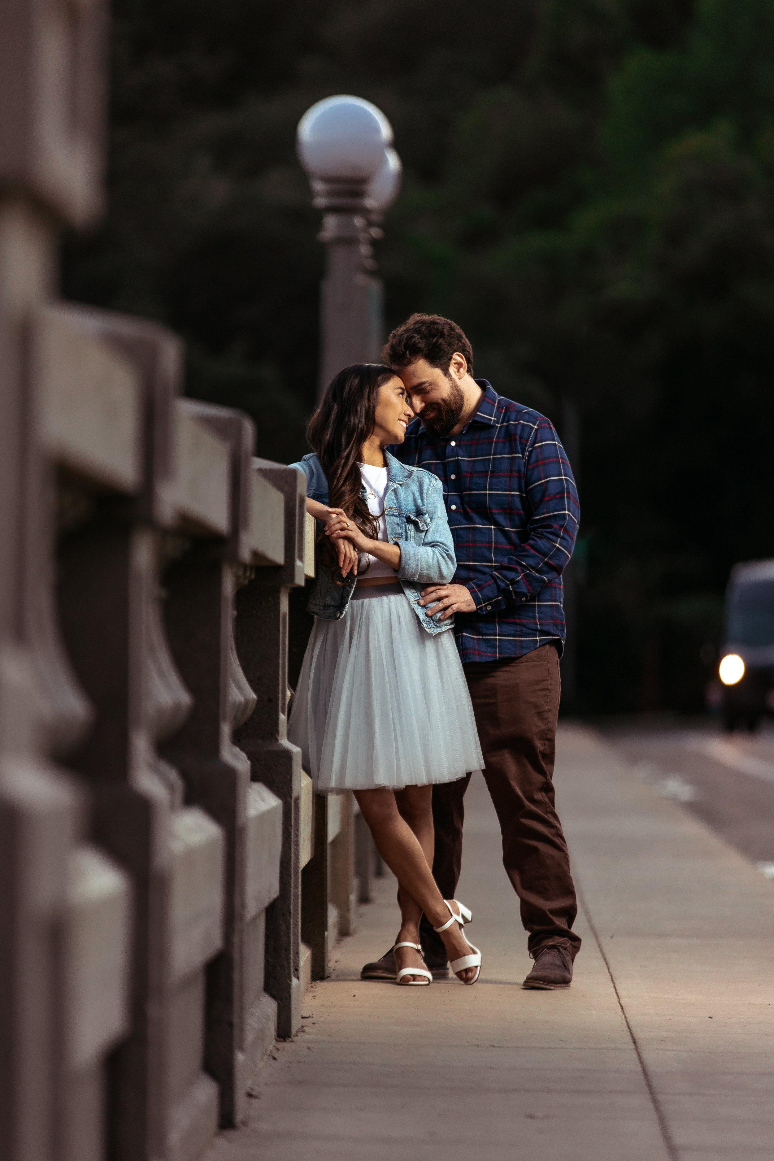 A couple standing close together on a sidewalk at dusk, smiling and leaning their foreheads together, with a streetlamp and car in the background.