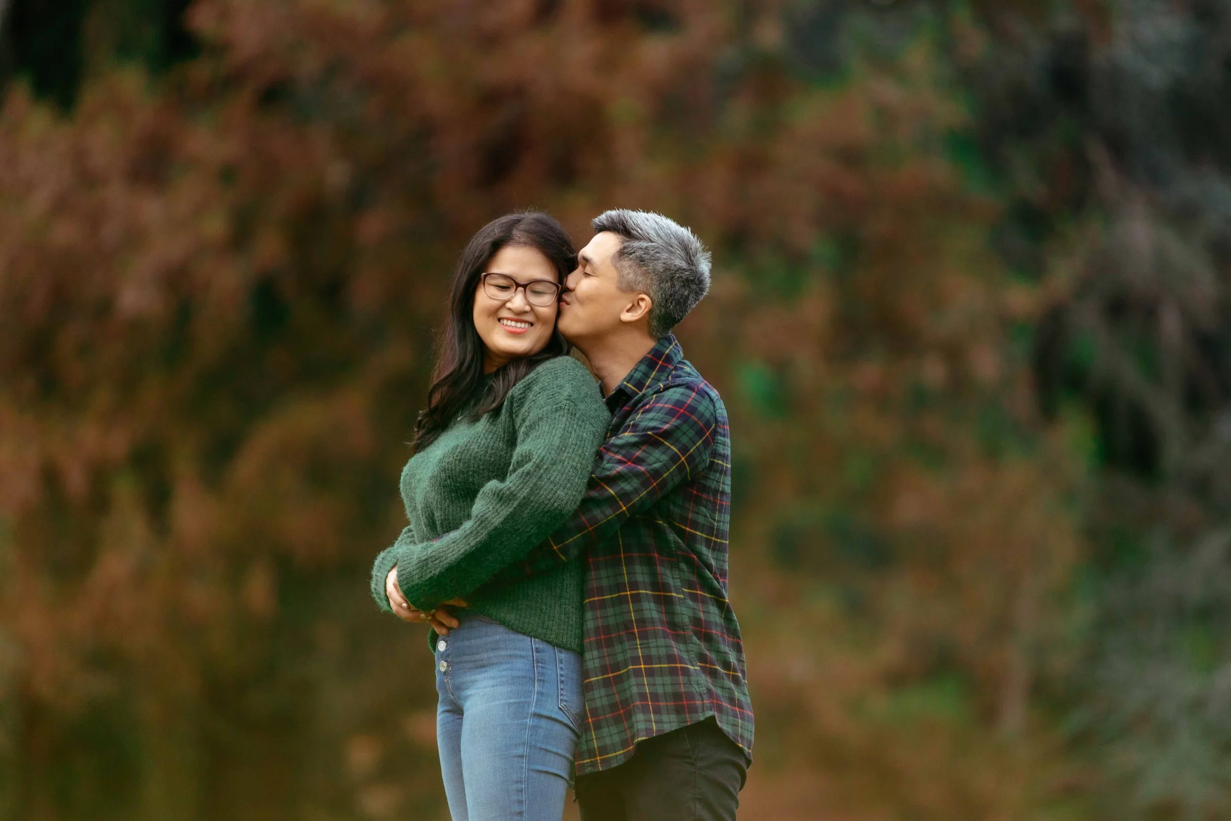 A couple embraces in a park with autumn trees, the man kissing the woman on the cheek as she smiles with eyes closed.