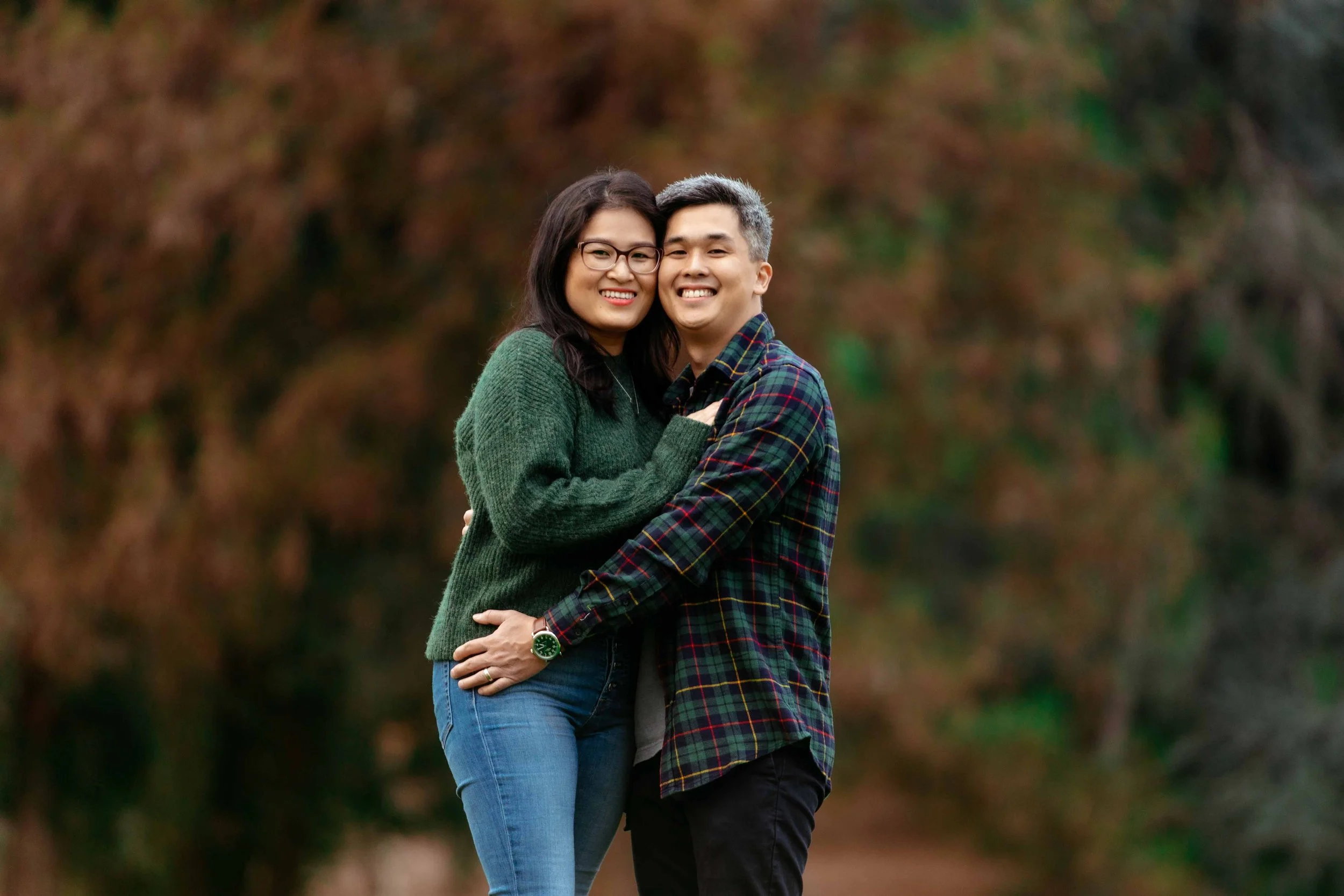 A smiling couple hugging outdoors with autumn foliage in the background.
