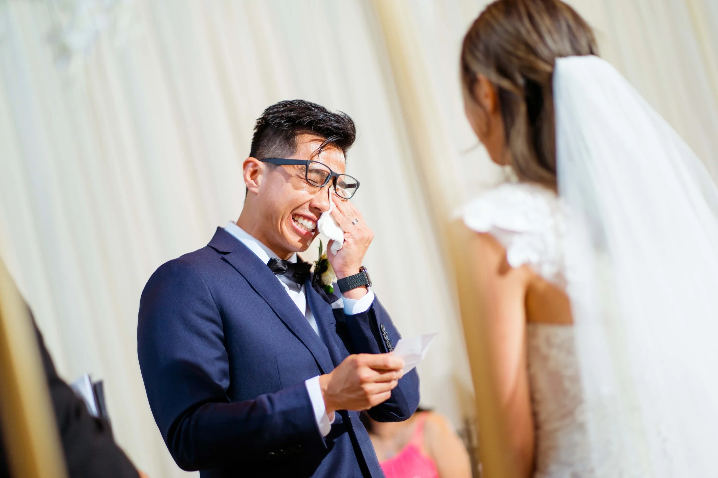 Groom in a navy suit and glasses wiping away tears while reading vows at his wedding, with a bride in a white lace dress and veil looking at him.