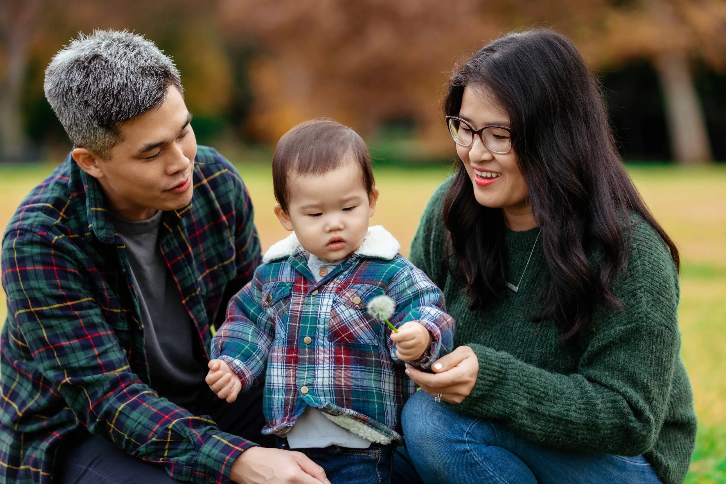 A family of three, including a father, a mother, and a young boy, sitting together outdoors in autumn. The mother is holding a dandelion puffball, and the young boy is looking at it while the father watches. They are all dressed in plaid and cozy clo
