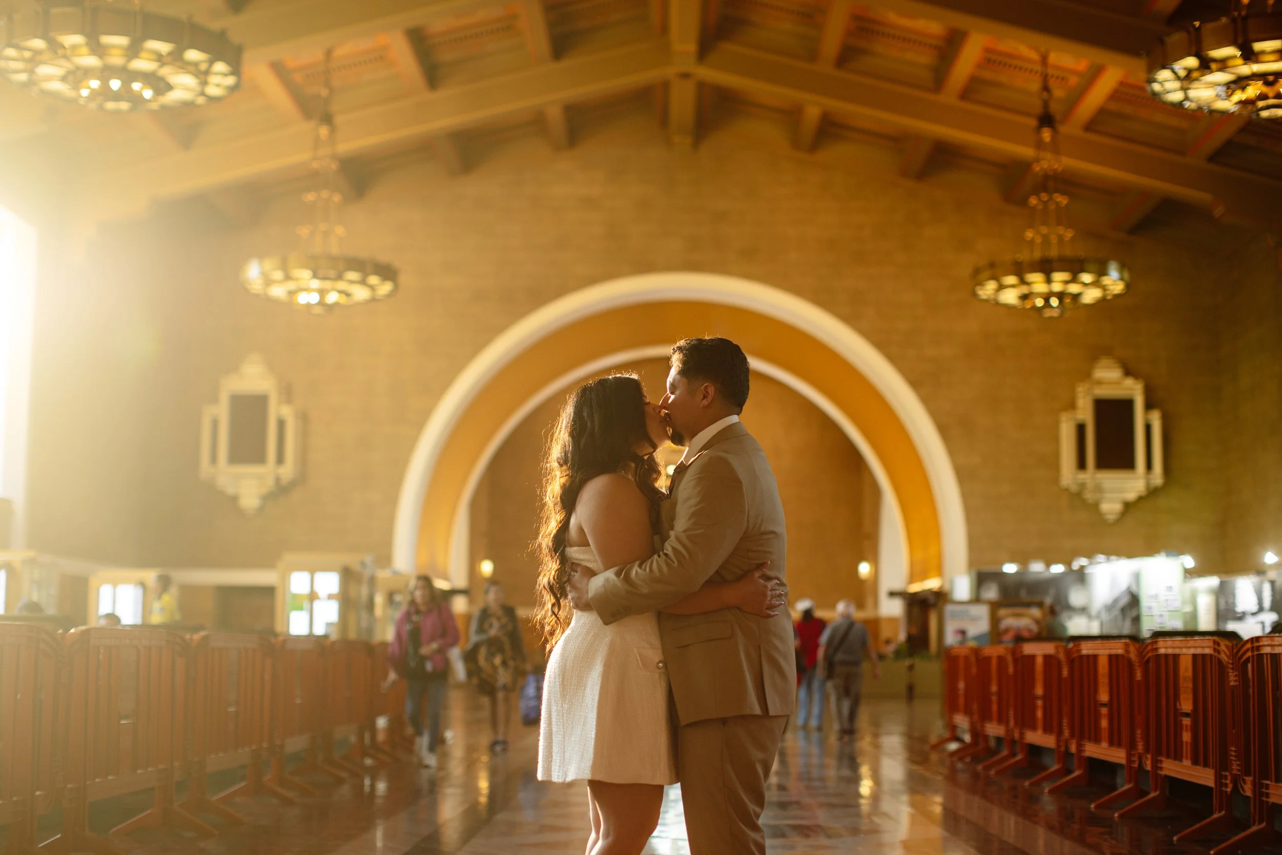 A couple kissing in an elegant, sunlit train station or terminal, with warm golden light, a high arched ceiling, and ticket barriers in the foreground.