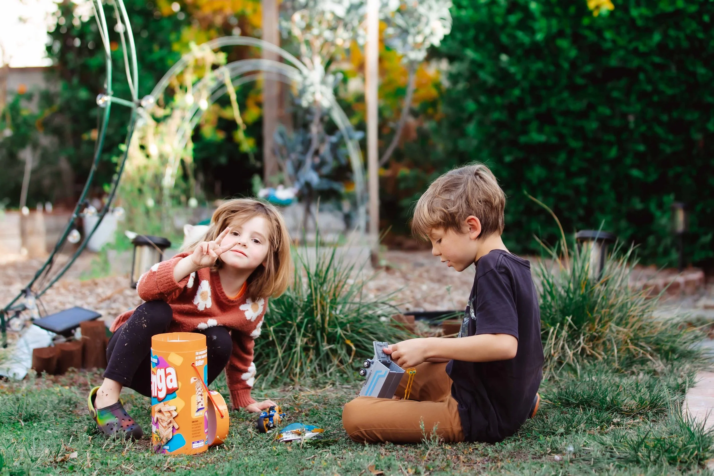 A young girl and boy playing outside on the grass, with the girl making a peace sign and the boy focused on a toy. There is a container of snacks or cookies nearby and a decorative metal arch in the background, along with lush greenery.