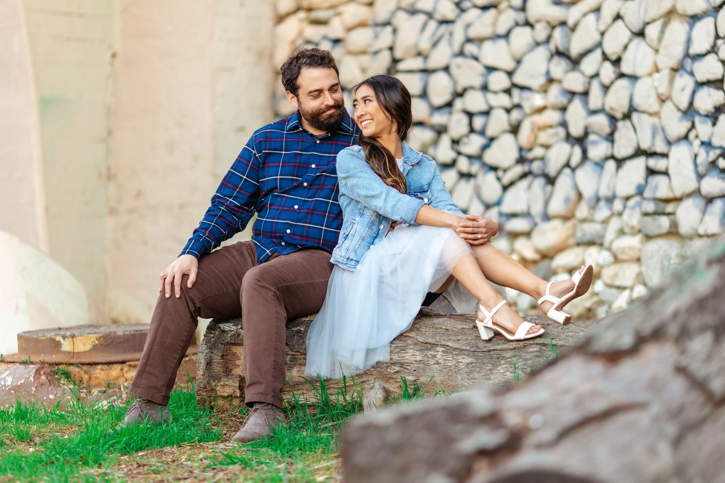 A couple sitting on a log outdoors, smiling and looking at each other, with a stone wall in the background.