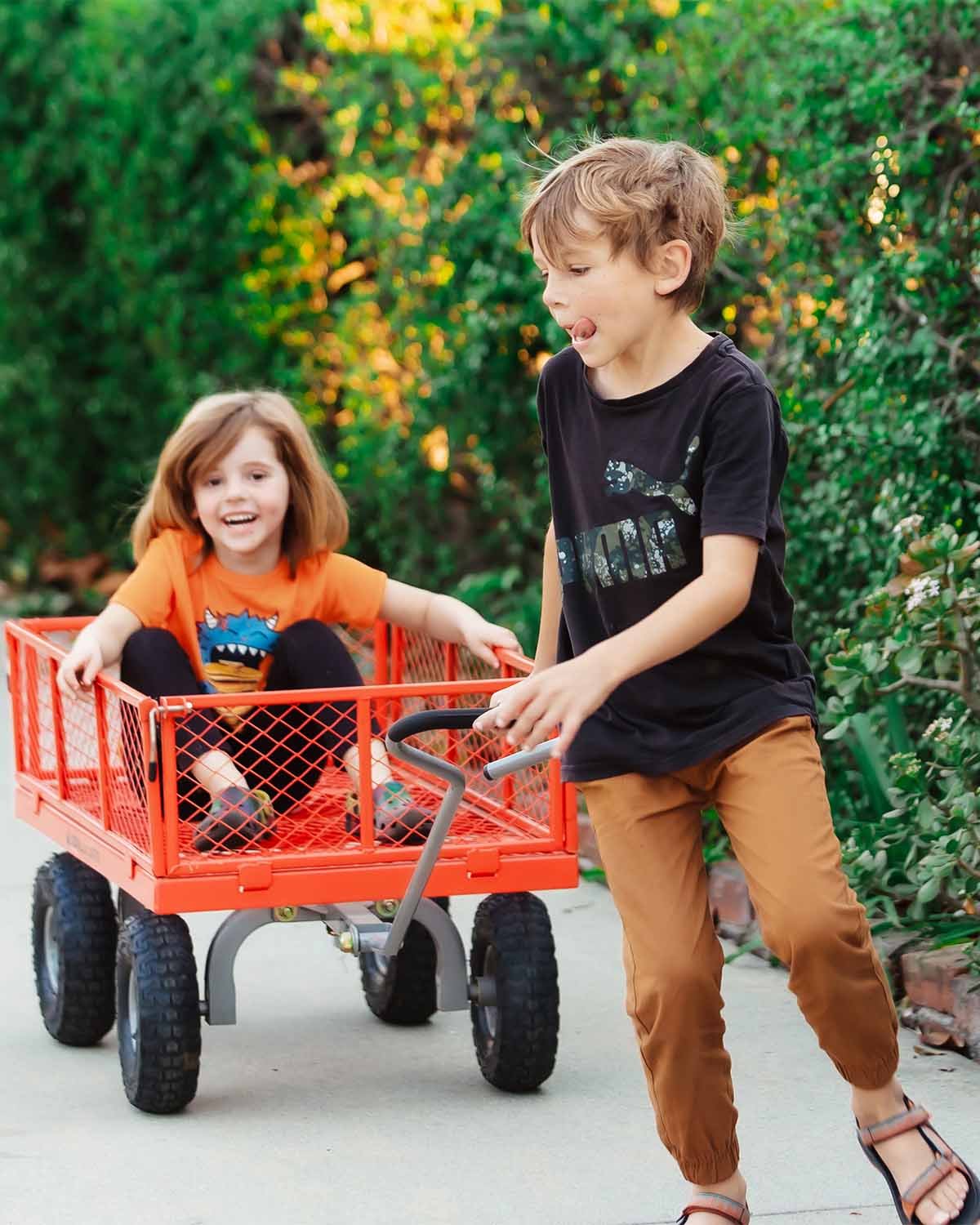 A young boy with brown hair pulling a red wagon with a smiling girl with red hair sitting inside, on a sidewalk surrounded by green bushes.