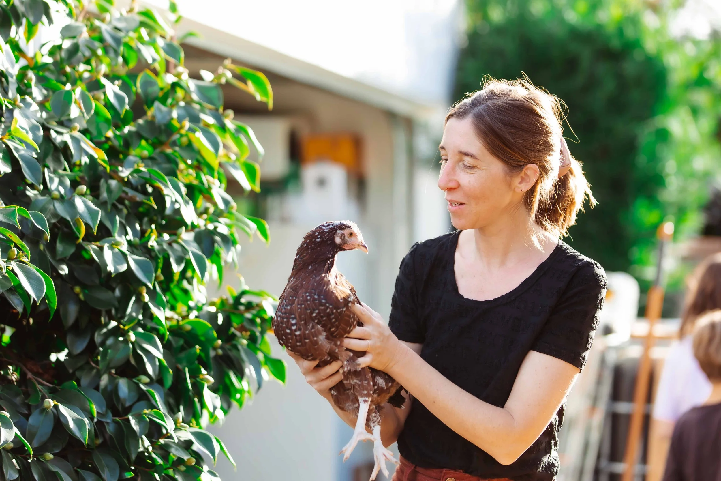 A woman holding a brown and white speckled chicken outside near green bushes, with a blurred background of trees and a house.