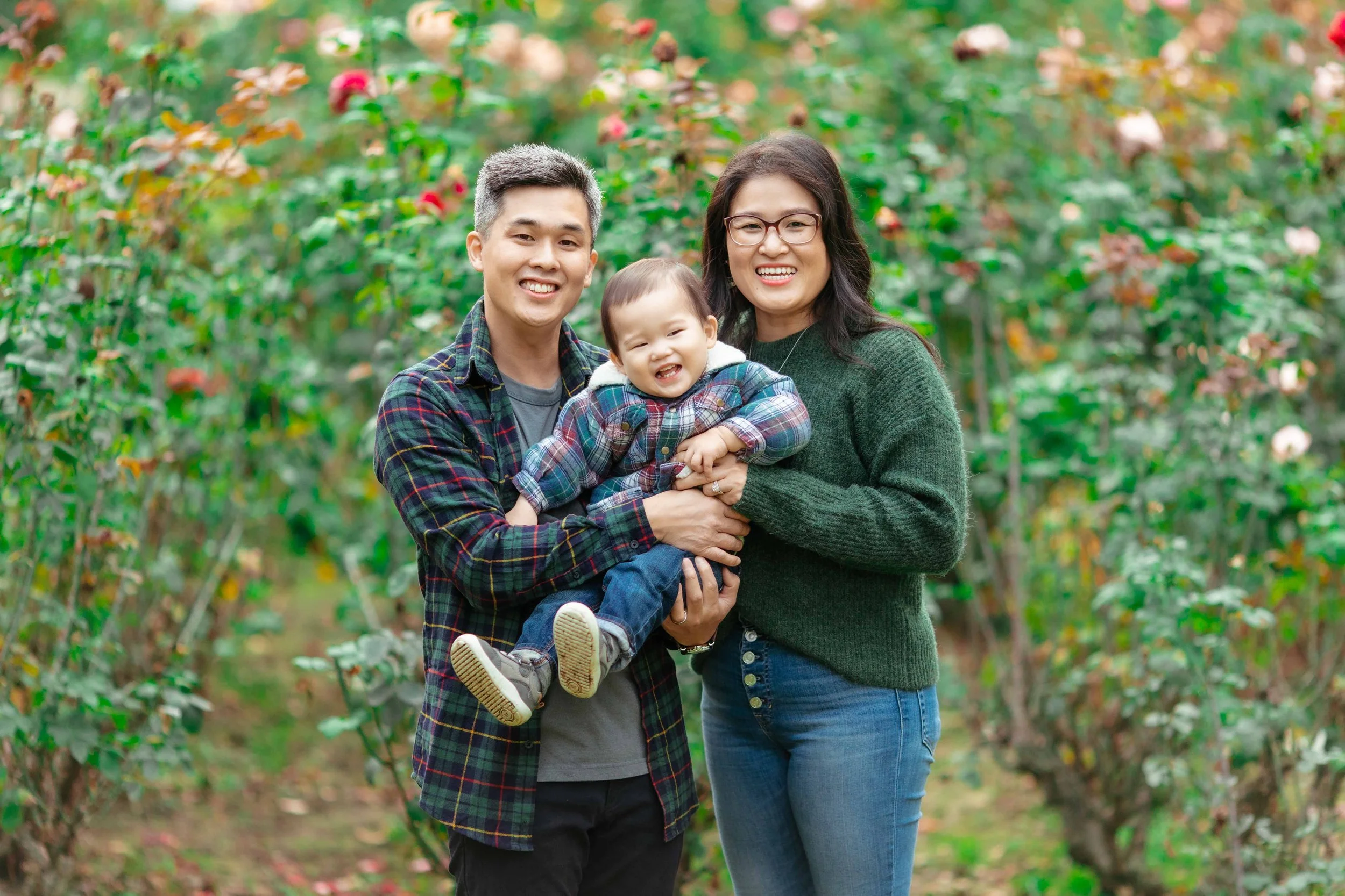 Family of three, a man, woman, and young child, smiling and standing in an orchard with trees bearing fruit.