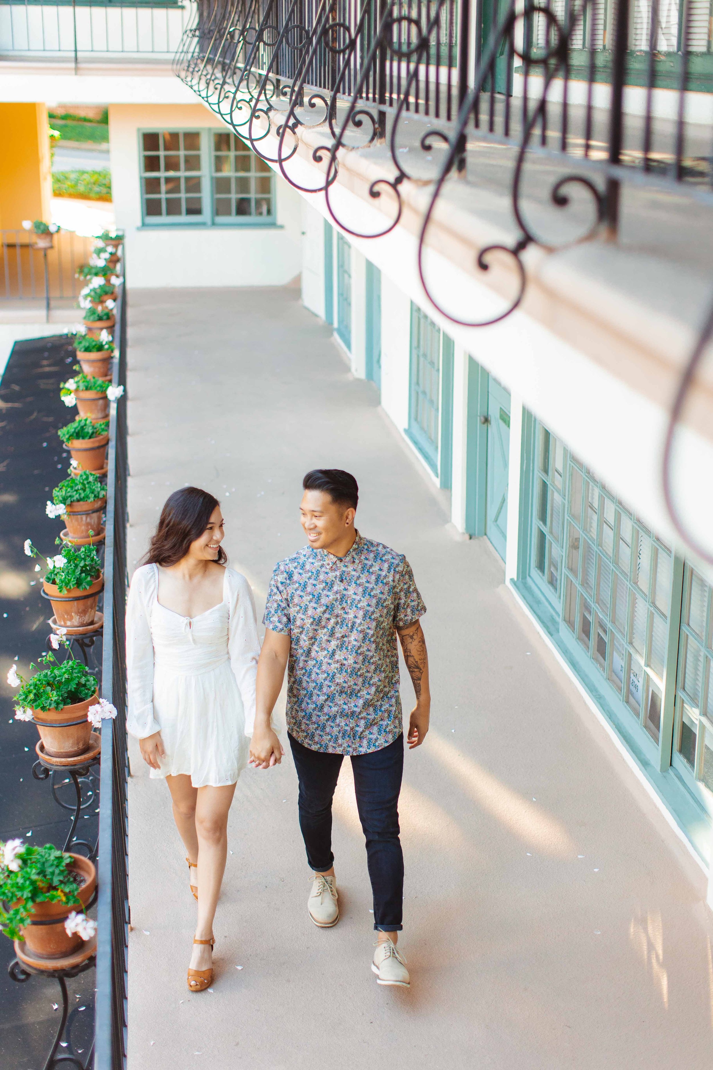A smiling couple holding hands and walking in an outdoor corridor with large windows, planters with flowers along the railing, and a colorful building exterior.