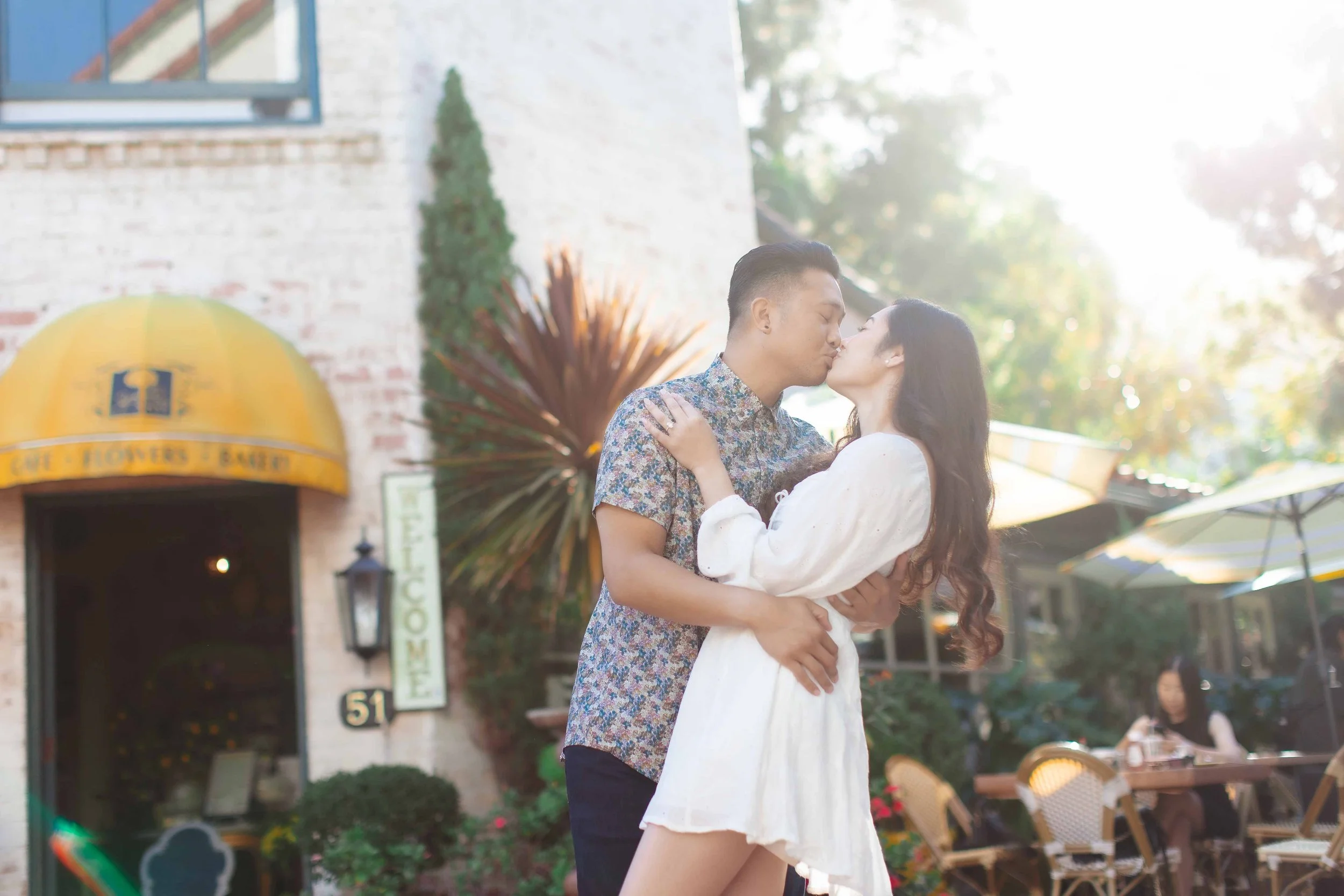 A couple sharing a kiss outdoors near a cafe, with patio tables, umbrellas, and a yellow awning in the background.