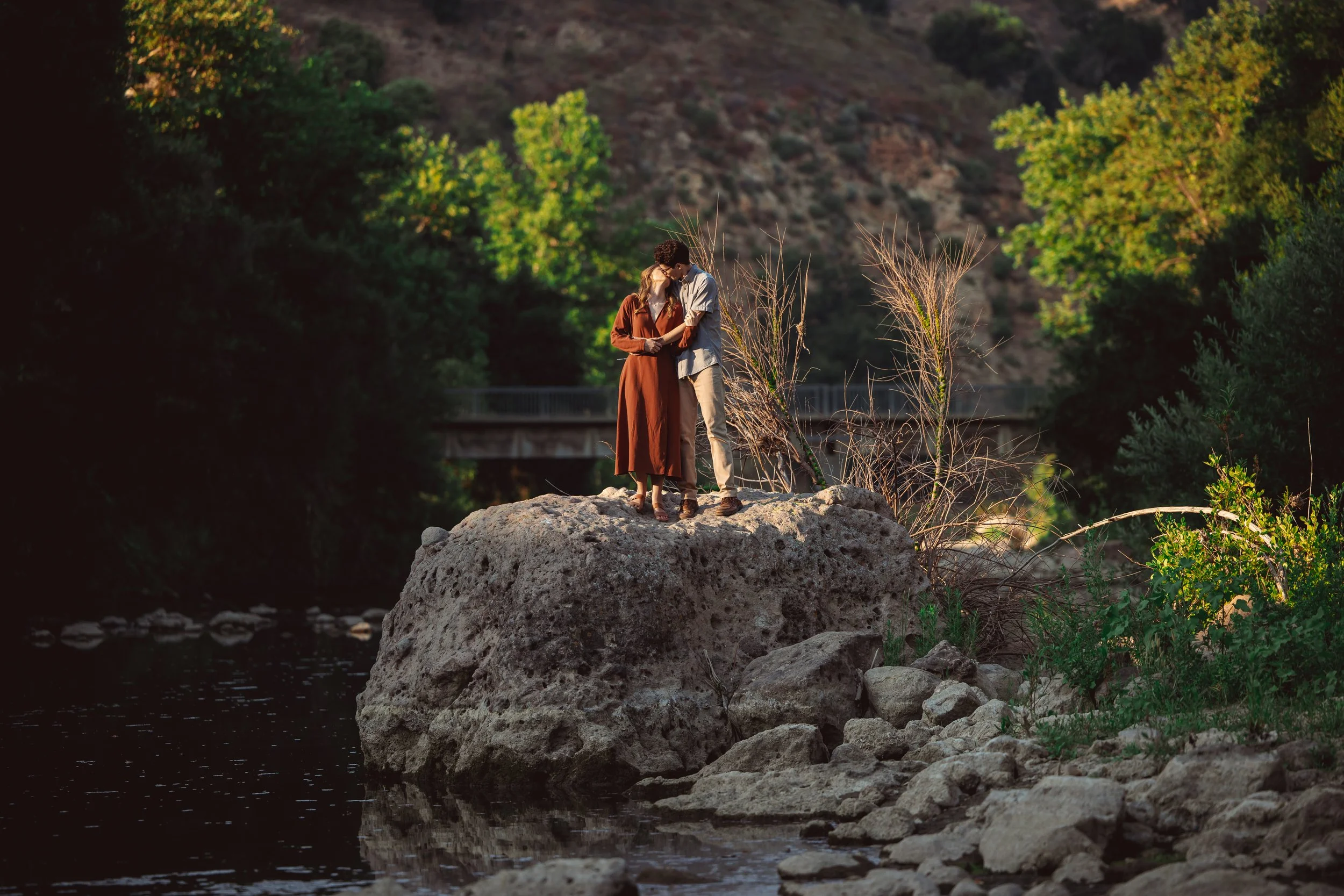 A couple standing on a large rock in a river, embracing each other with trees and mountains in the background during sunset.