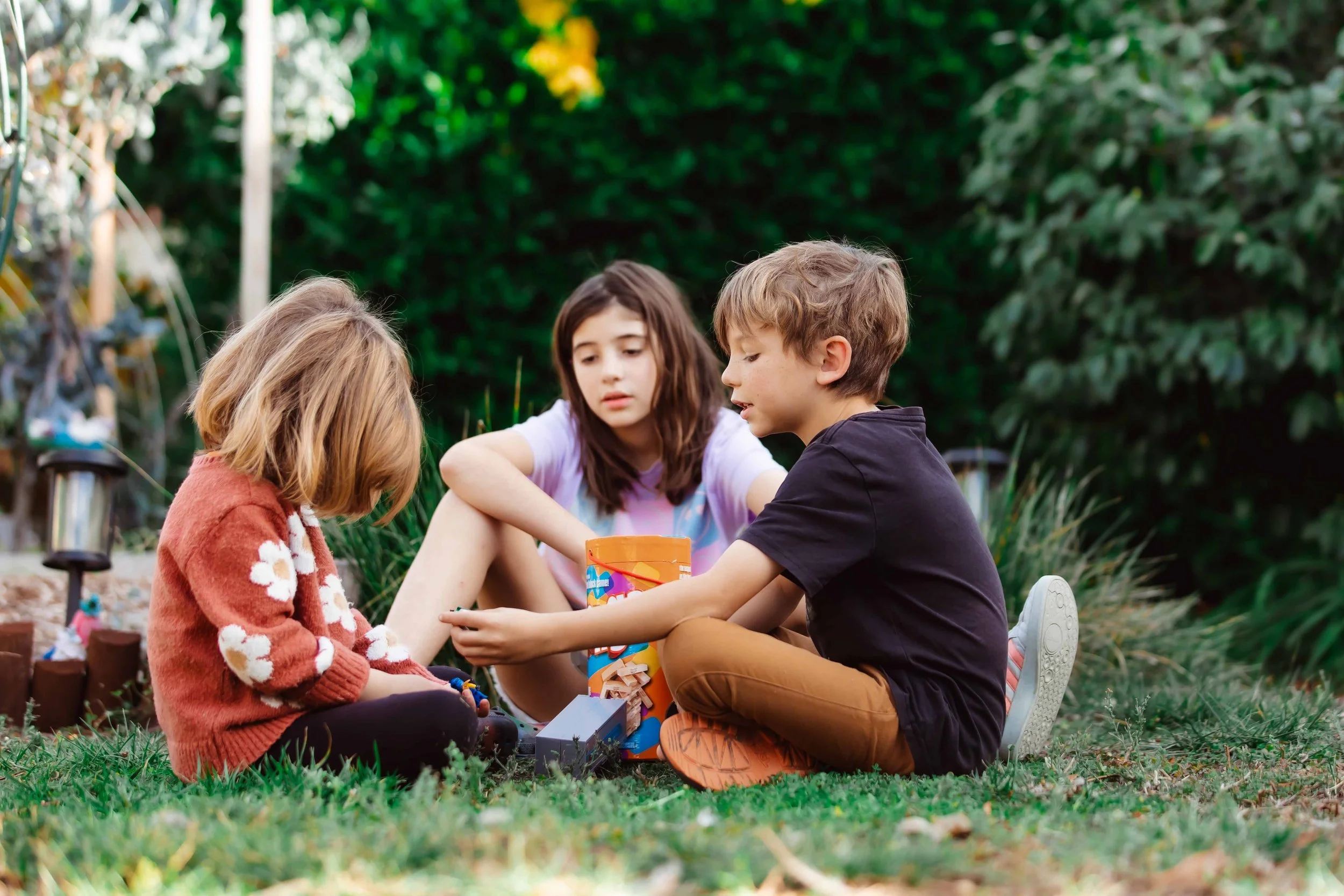 Three children sitting outdoors on grass, playing with toys and a bucket.