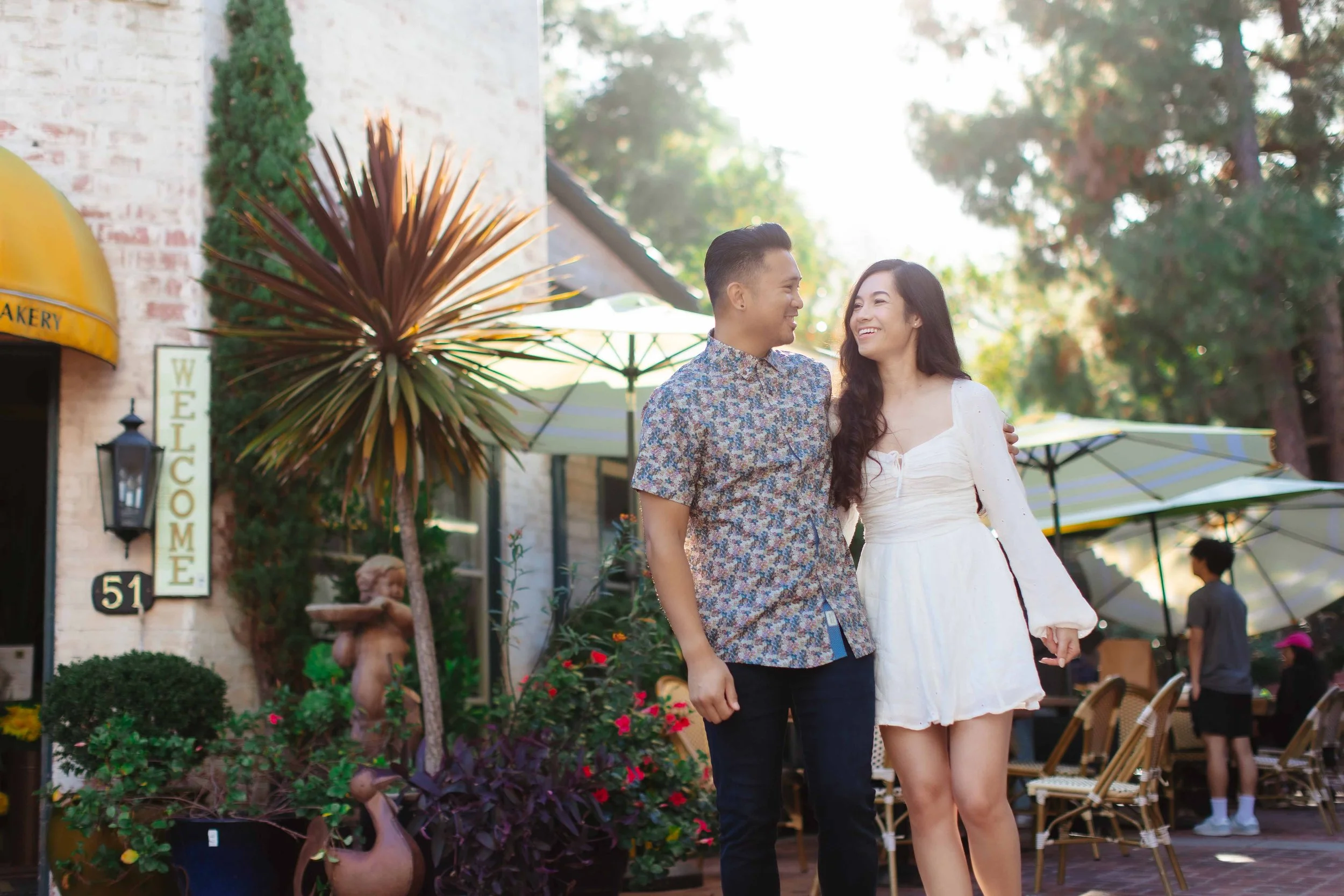 A young couple walking arm in arm, smiling at each other, in an outdoor shopping or dining area with umbrellas, plants, and a 'Welcome' sign on a building.