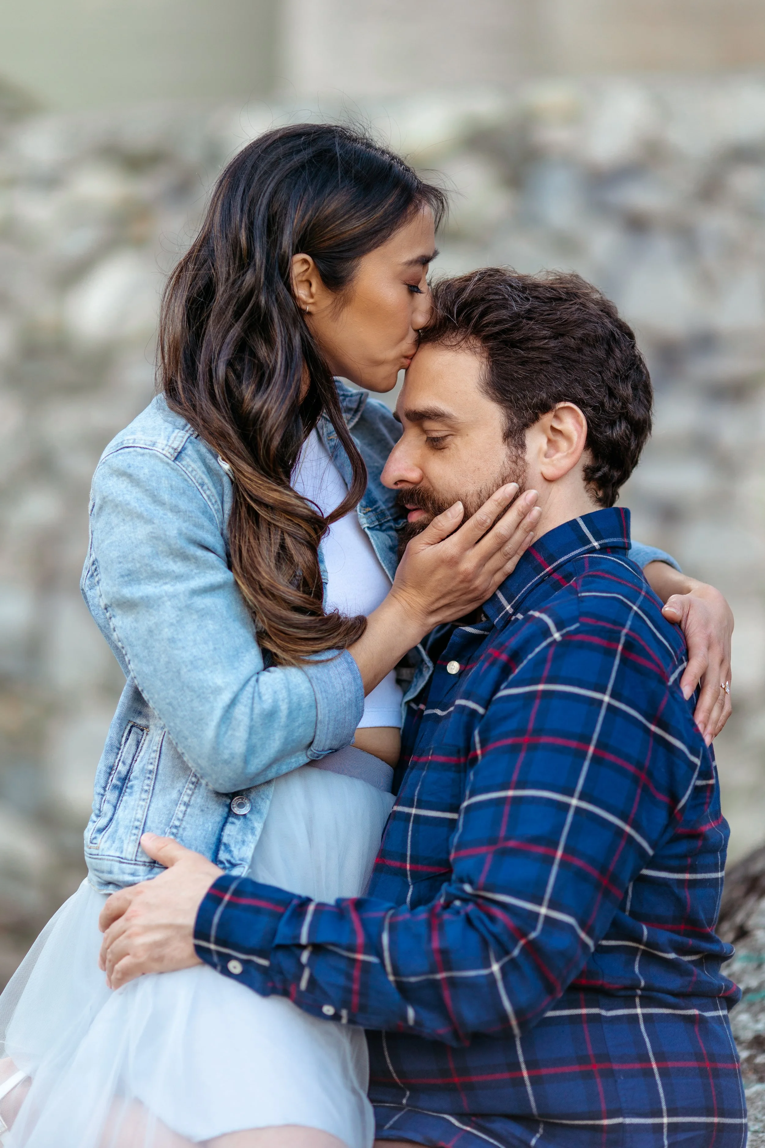 A woman kissing a man on the forehead while holding his face and his hand resting on her waist, outdoors with a rocky background.