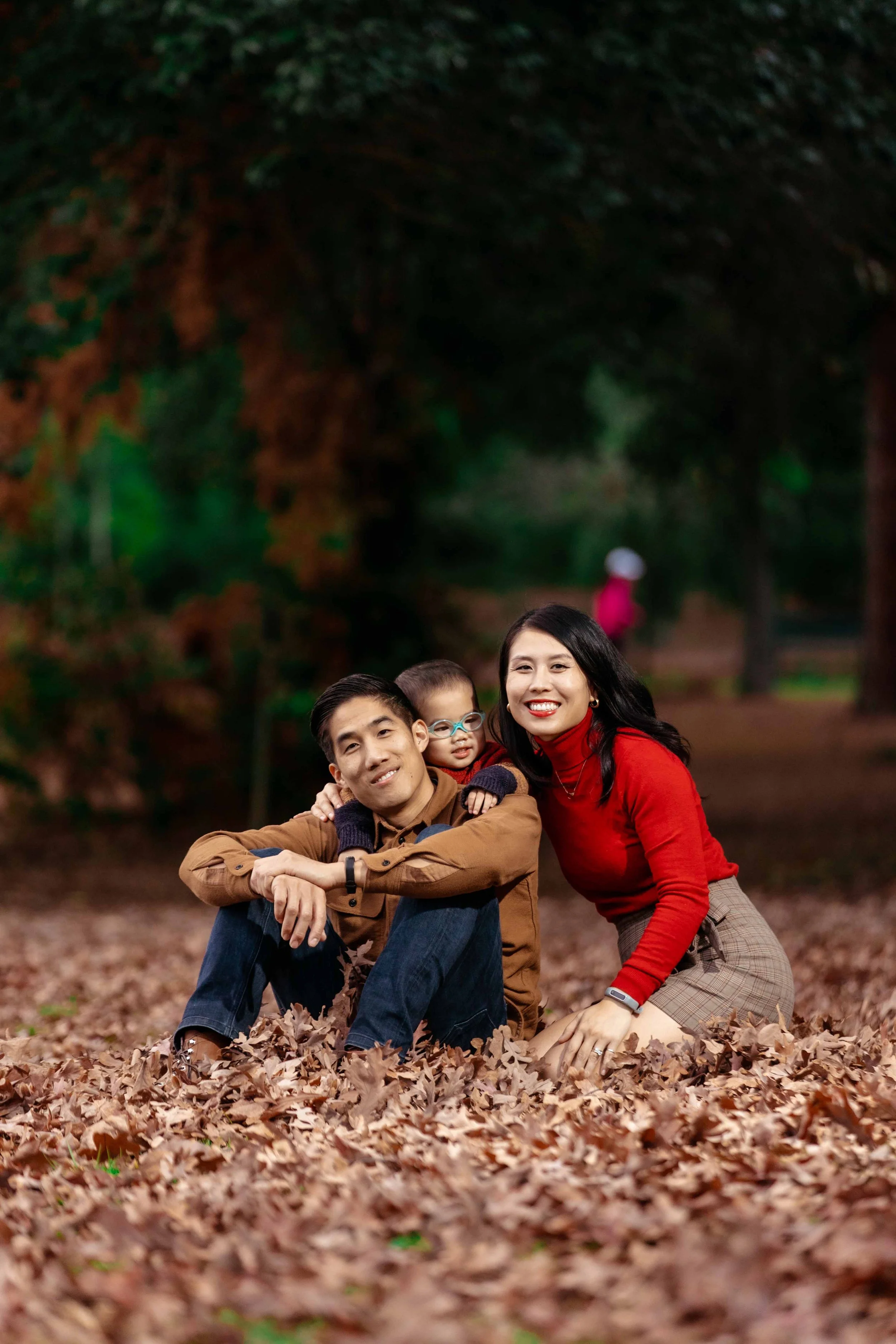 A family of three, a man, a woman, and a child, sitting on a bed of fallen leaves in a park during autumn, smiling and enjoying the outdoor scenery.