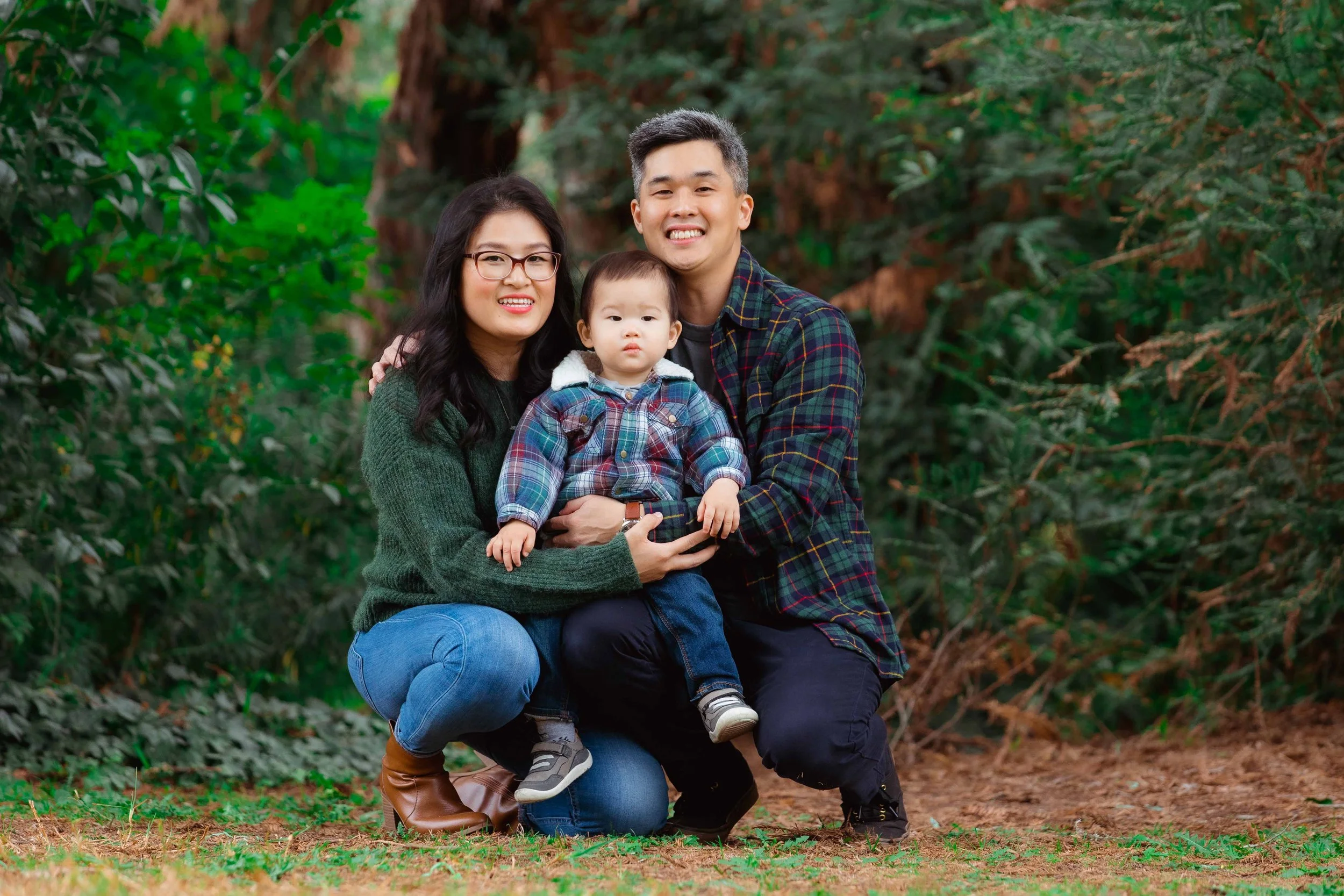 A family of three posing outdoors in a wooded area. The mother and father are kneeling, holding their young son between them. The mother has dark hair, glasses, and is wearing a green sweater. The father has short dark hair and is wearing a plaid shi