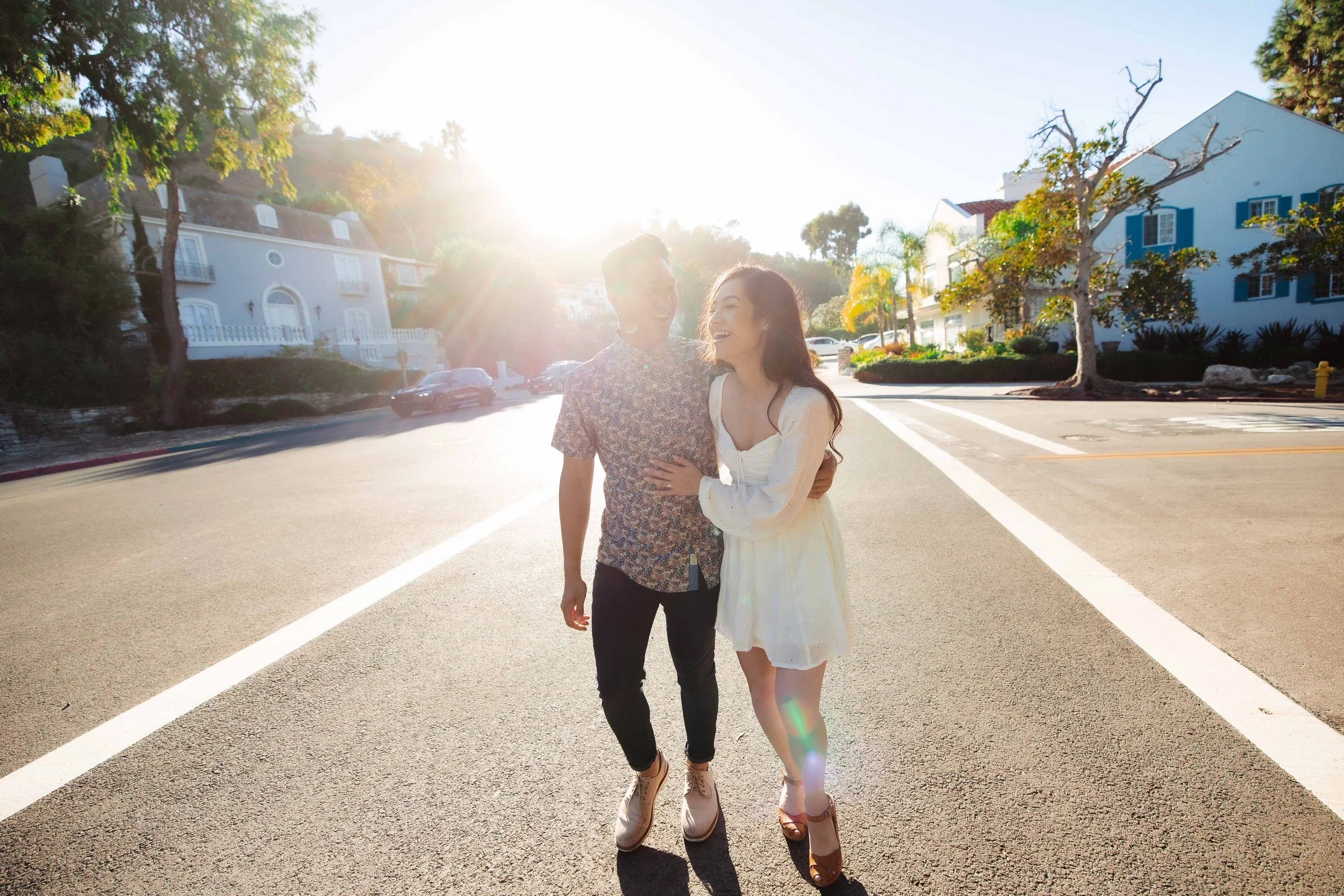 A young couple laughing and holding each other on a sunny suburban street with colorful houses and trees.