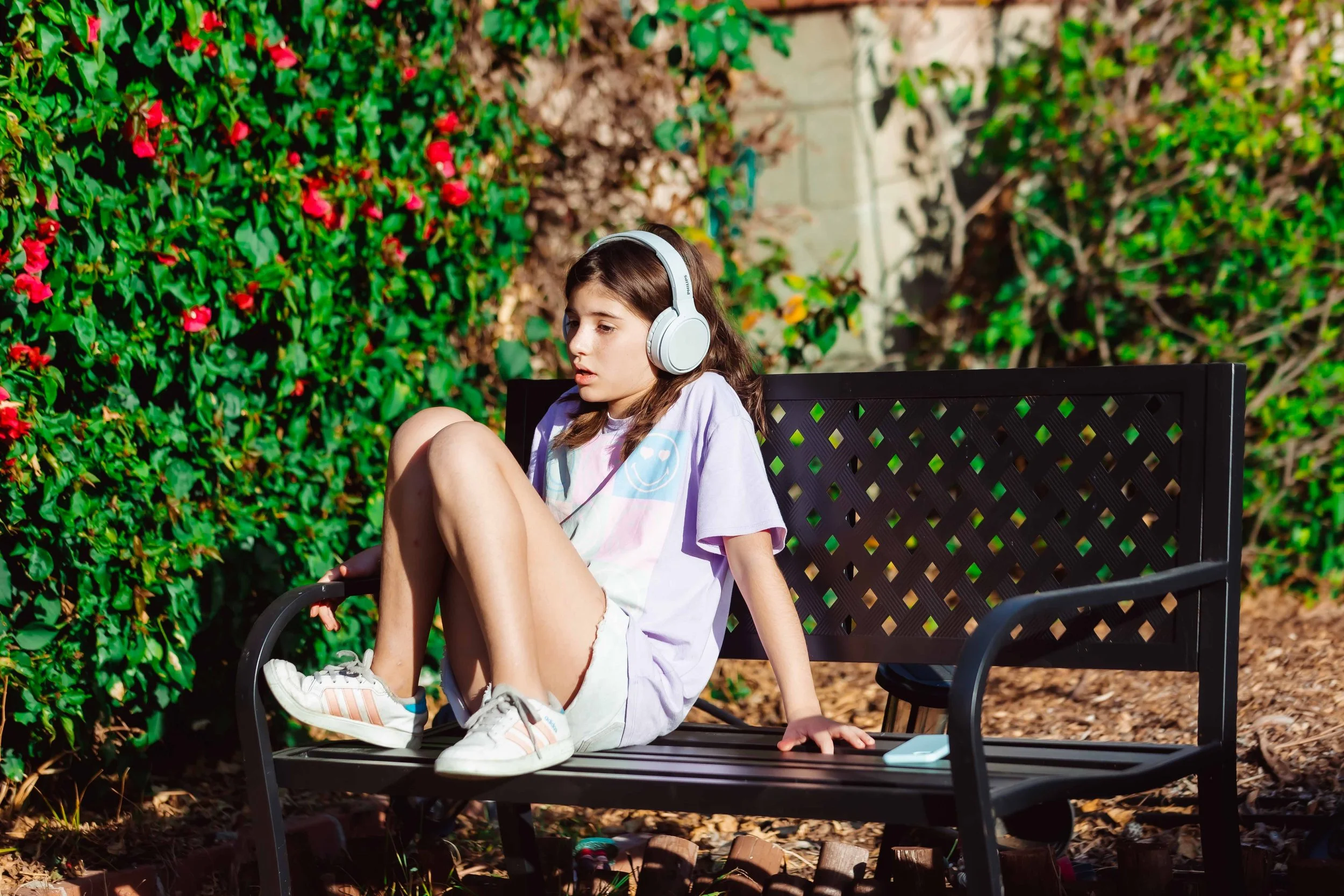 A young girl sitting on a black outdoor bench, wearing headphones, a pastel tie-dye t-shirt, shorts, and sneakers, in a garden with bushes and flowers.