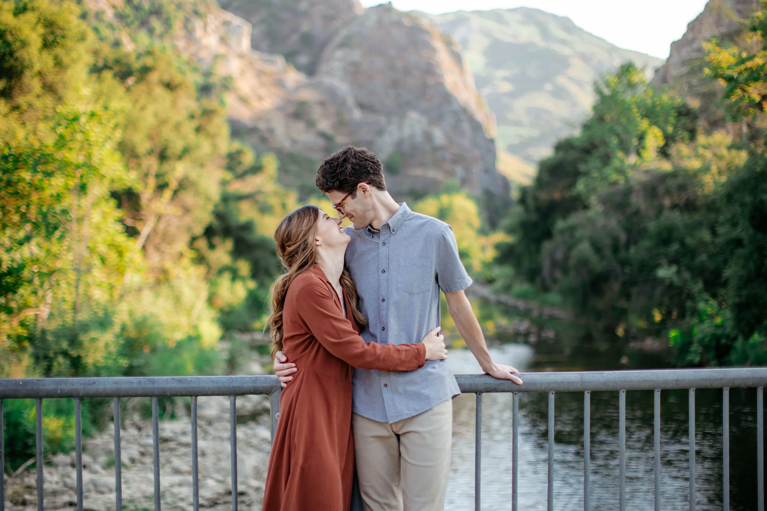 A young couple standing close together on a bridge near a river, surrounded by green trees and mountains, smiling at each other.