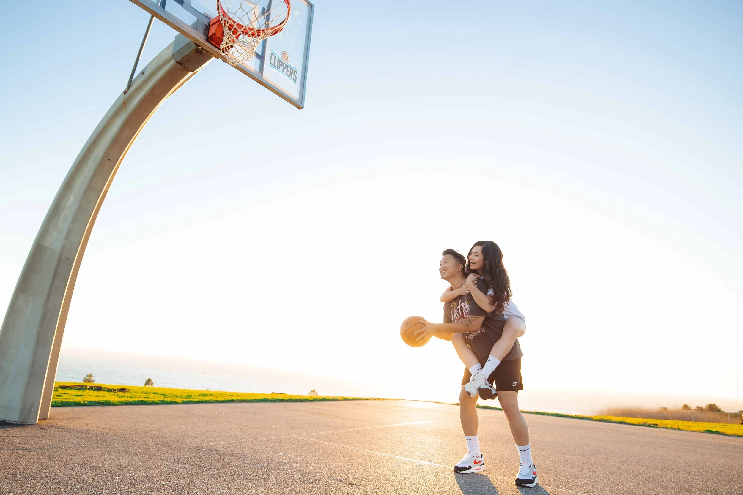 A young man giving a piggyback ride to a young woman on an outdoor basketball court during sunset, with the basketball hoop in the background.