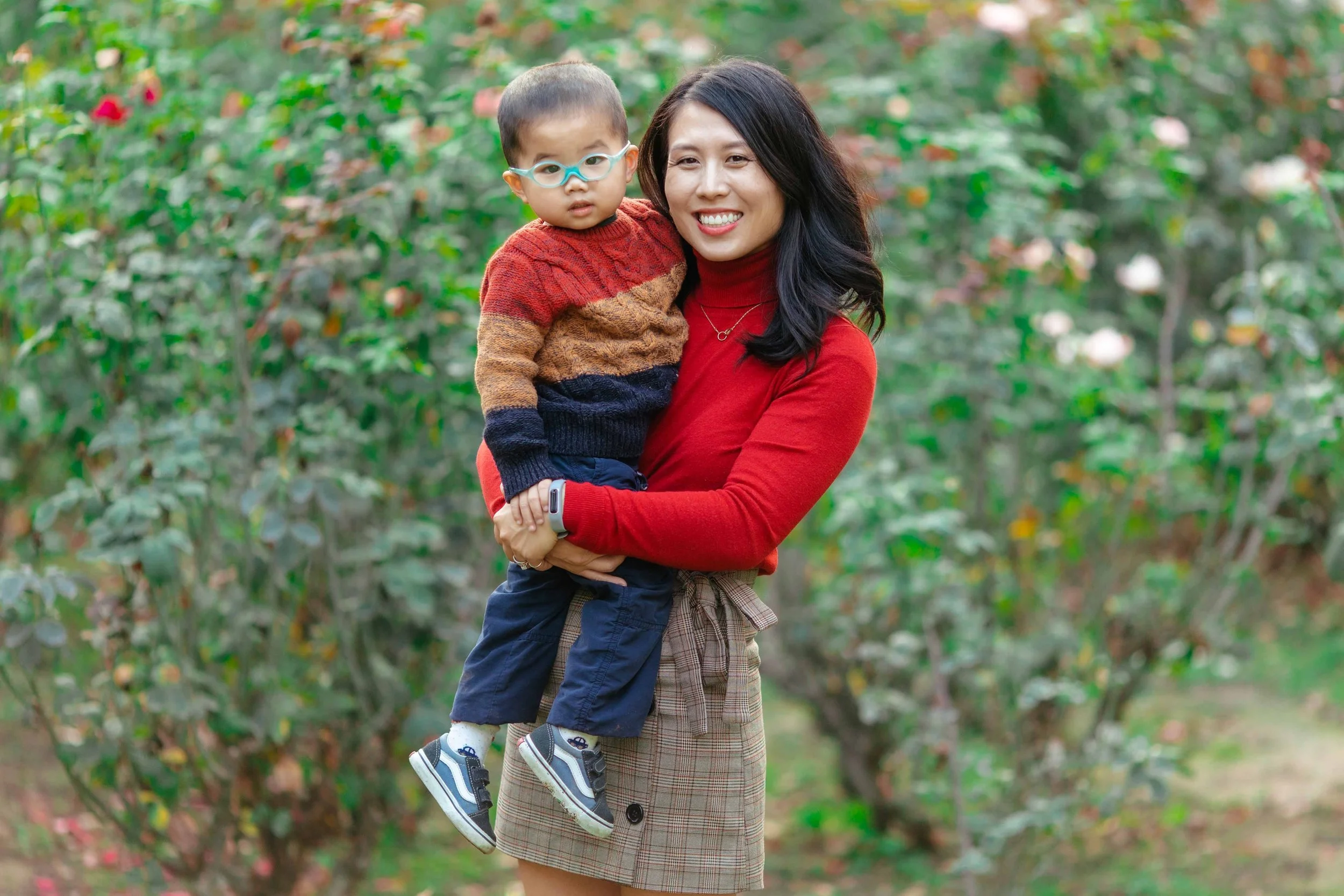 A woman in a red turtleneck holding a young boy outdoors surrounded by greenery and blooming pink flowers.