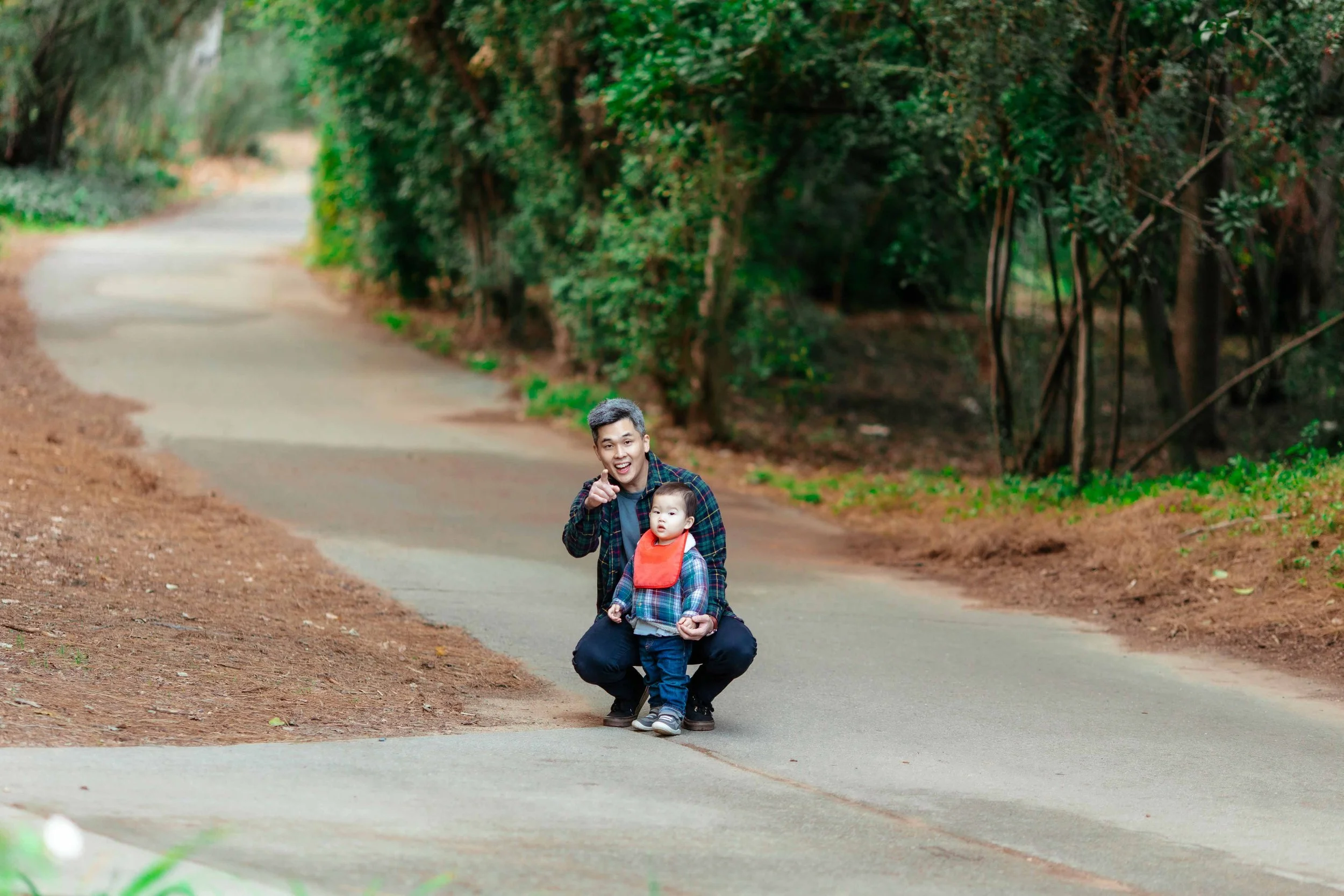 A man and a young child are on a paved path in a wooded area. The man is squatting, smiling, and pointing at the camera, while the child stands in front of him wearing a red bib and plaid shirt.