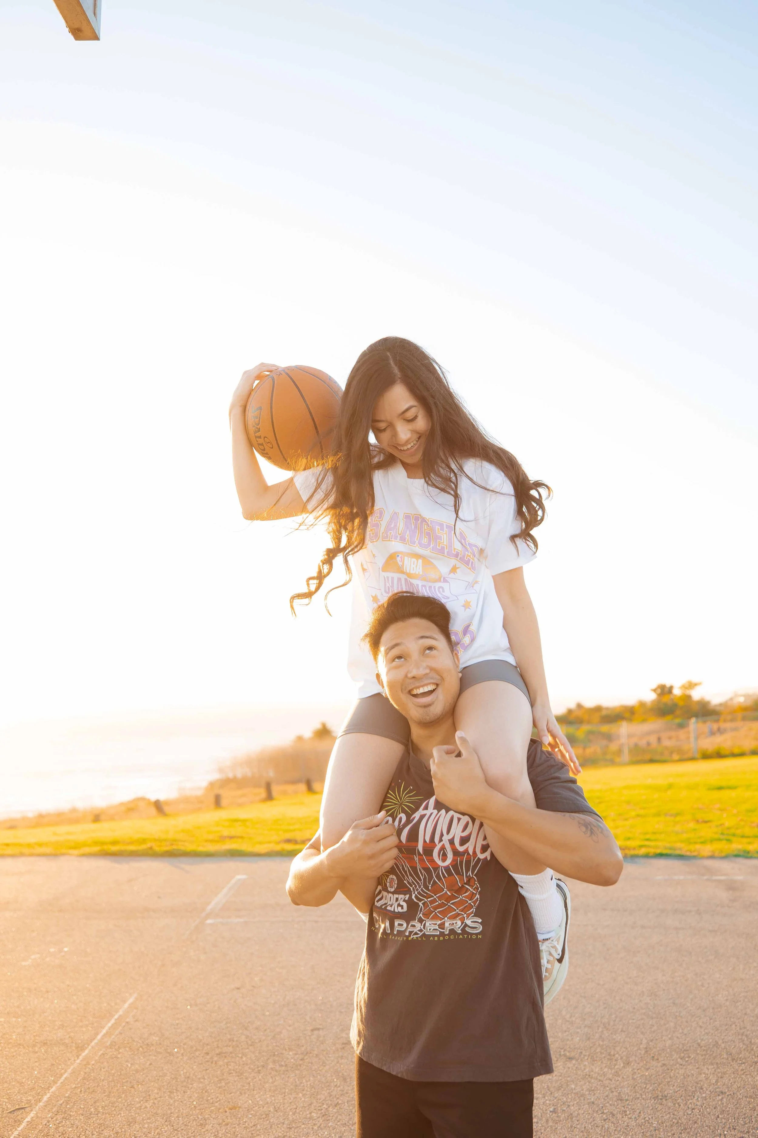 A joyful young man is holding a young woman on his shoulders on an outdoor basketball court during sunset. The woman is holding a basketball, smiling and looking down at the man. They are casually dressed and the background shows a grassy field and a