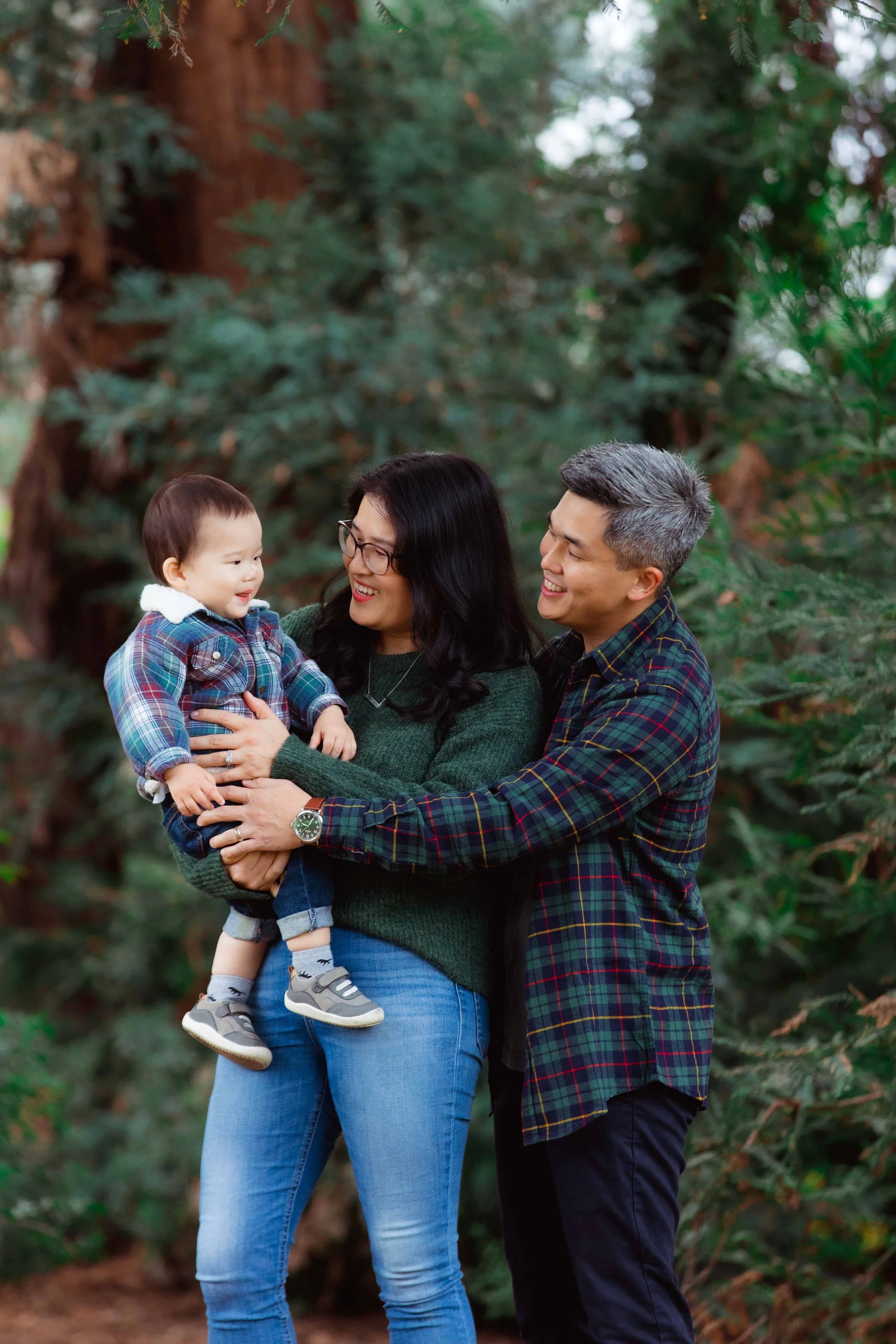 A happy family of three at a forest, with a woman holding a toddler boy and a man reaching out to him, all smiling.