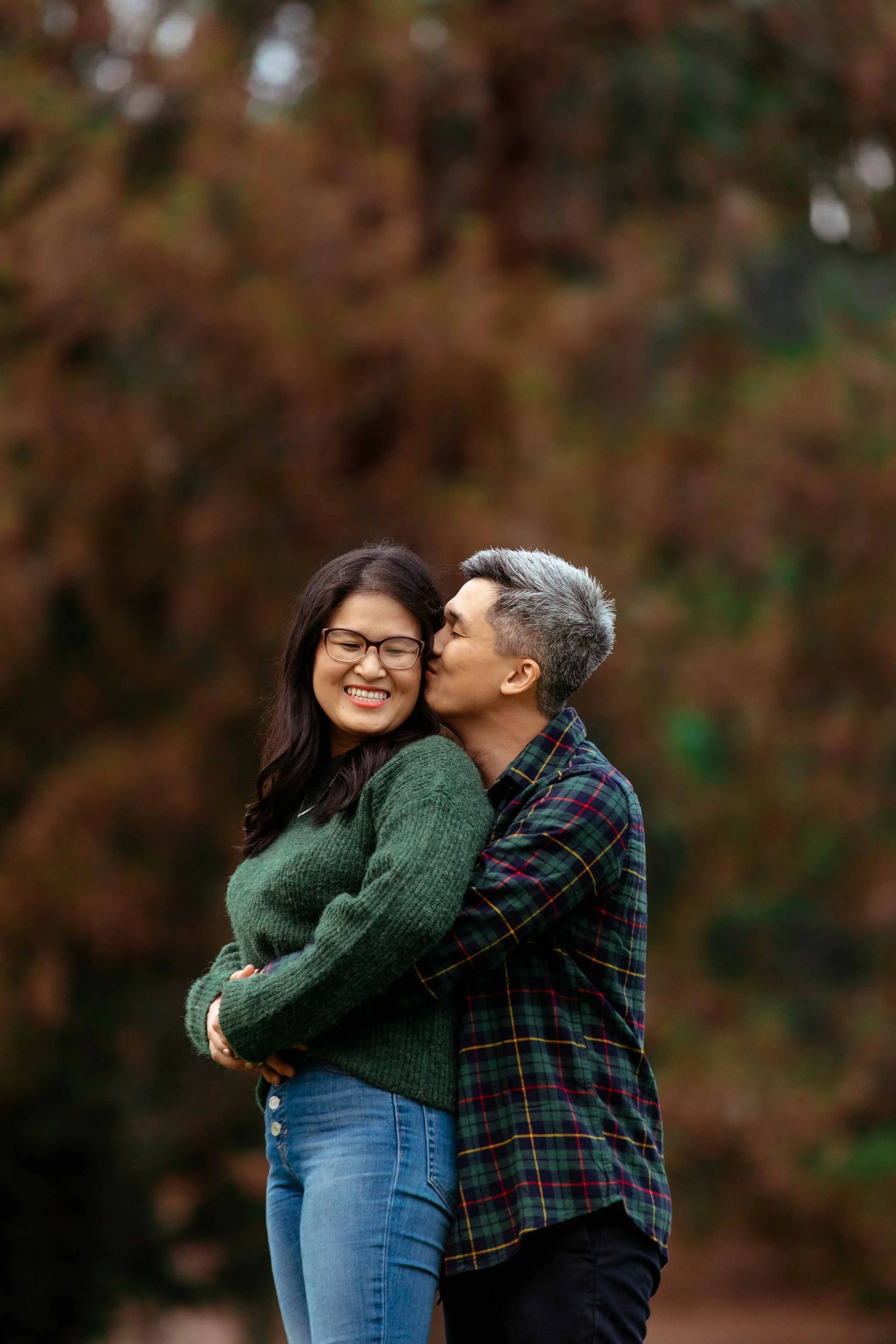 A man giving a woman a kiss on the cheek outdoors during autumn, with colorful fall foliage in the background. The woman is smiling and wearing glasses and a green sweater, while the man is dressed in a plaid shirt.