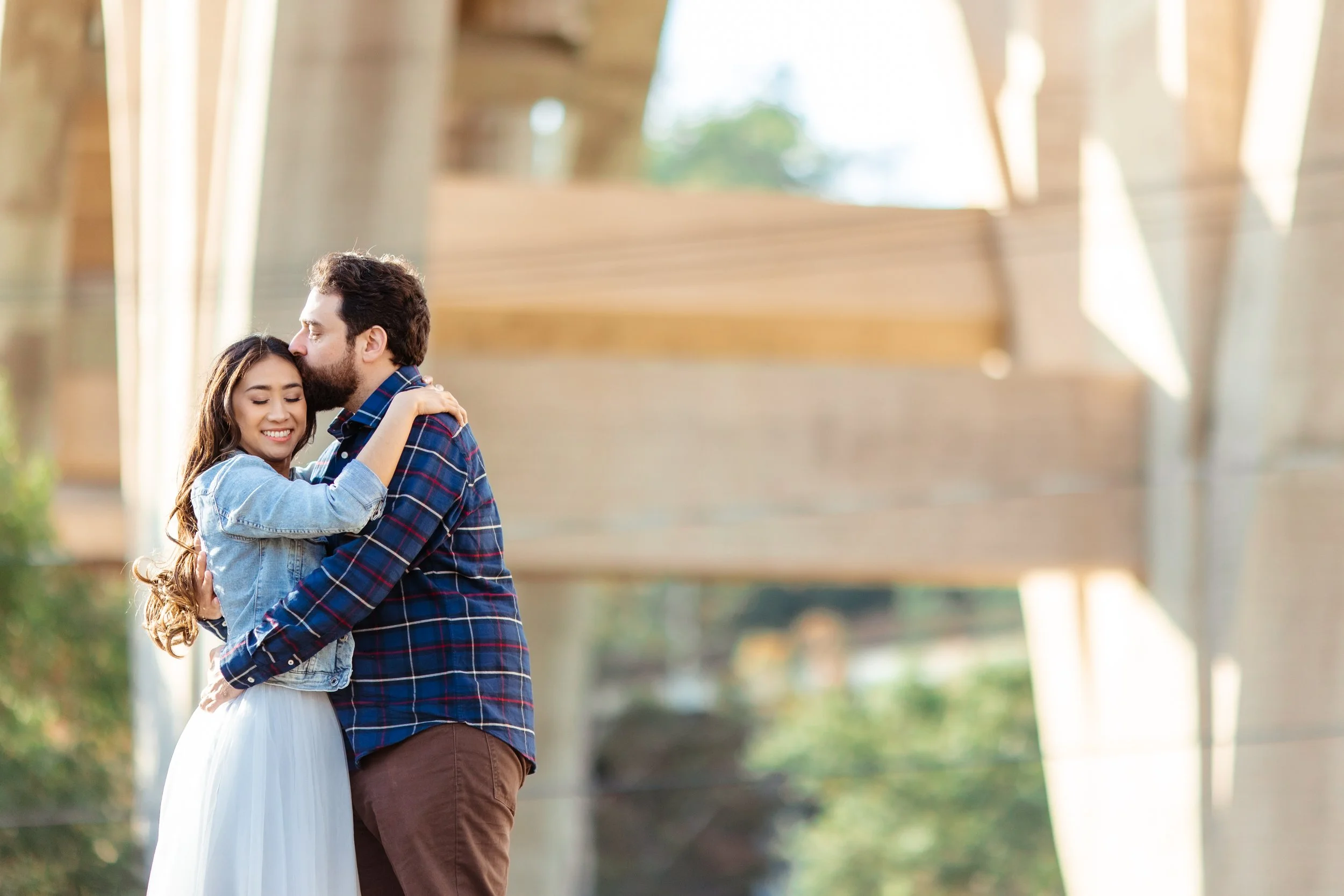 A young woman and a man hugging outdoors under a bridge with trees in the background.