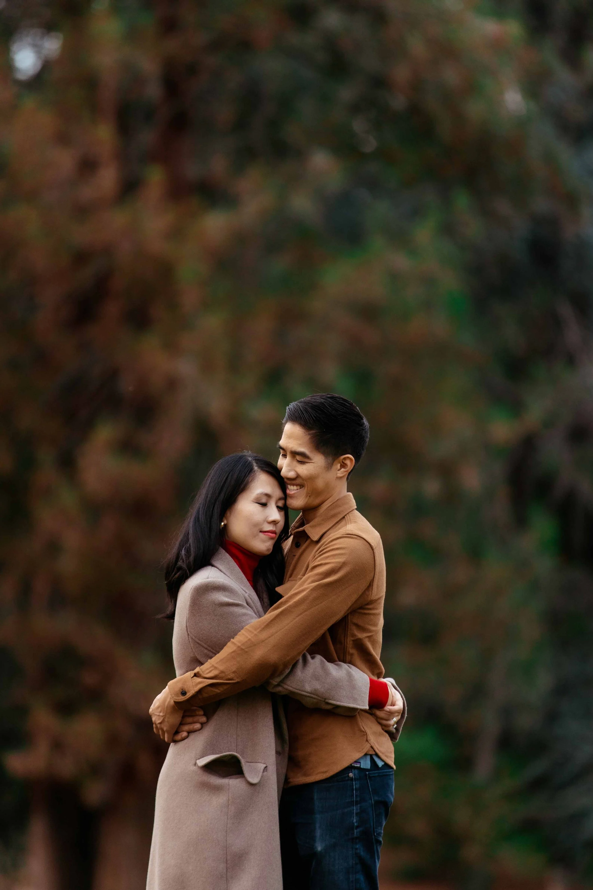 A couple hugging outdoors in front of blurred trees.