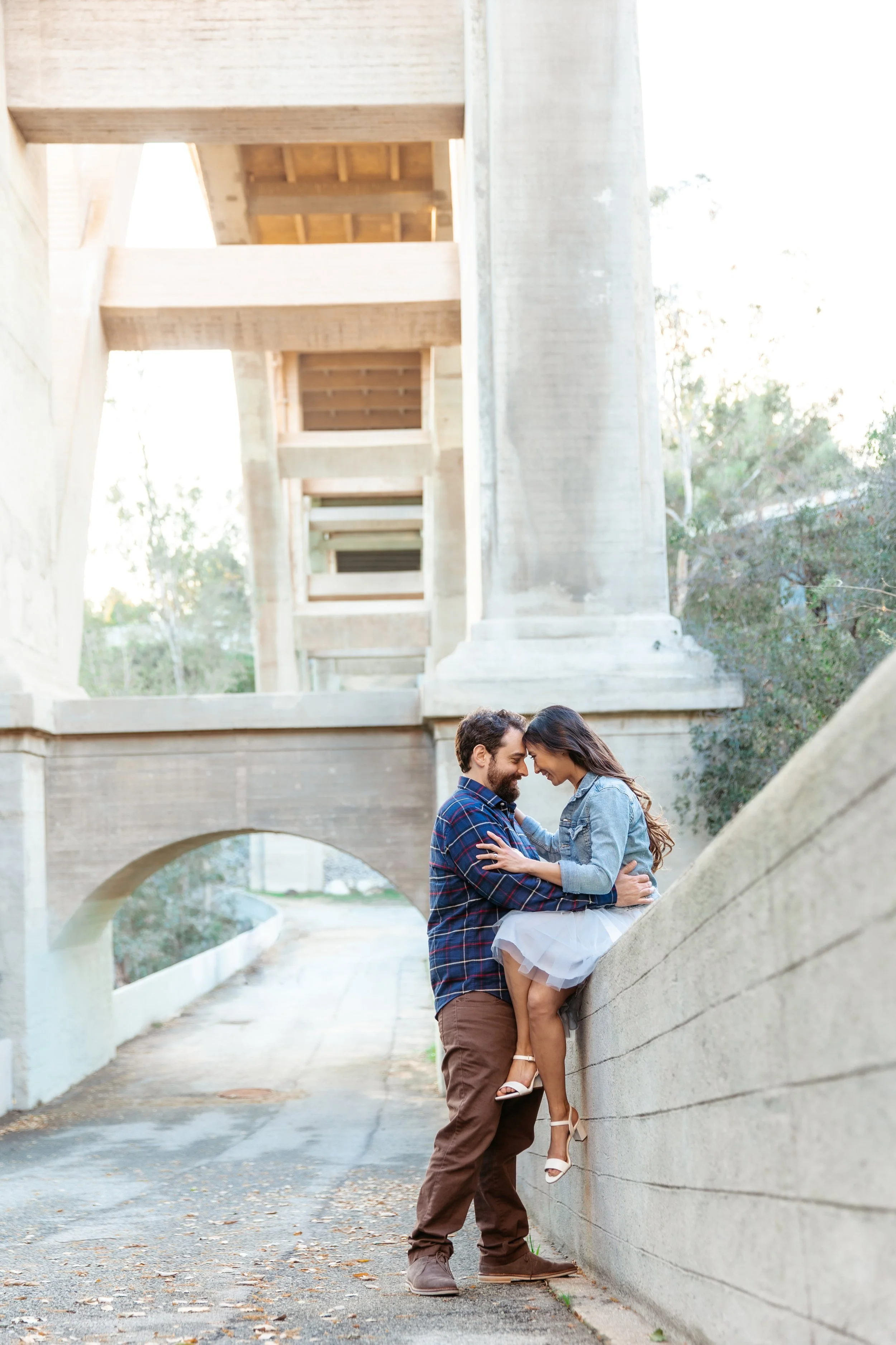 A couple sharing an intimate moment with foreheads touching under a bridge, with the woman sitting on a ledge and the man standing.