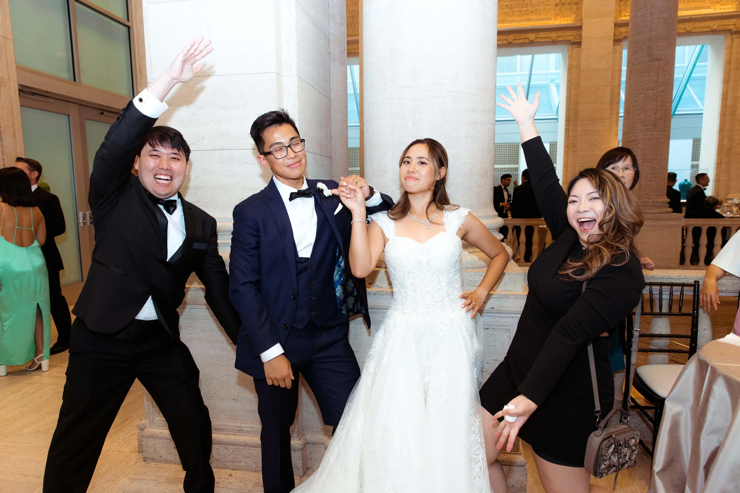 People celebrating at a wedding reception, with a bride in a white wedding dress and three friends, two men in tuxedos and one woman in black, posing together with joyful expressions.