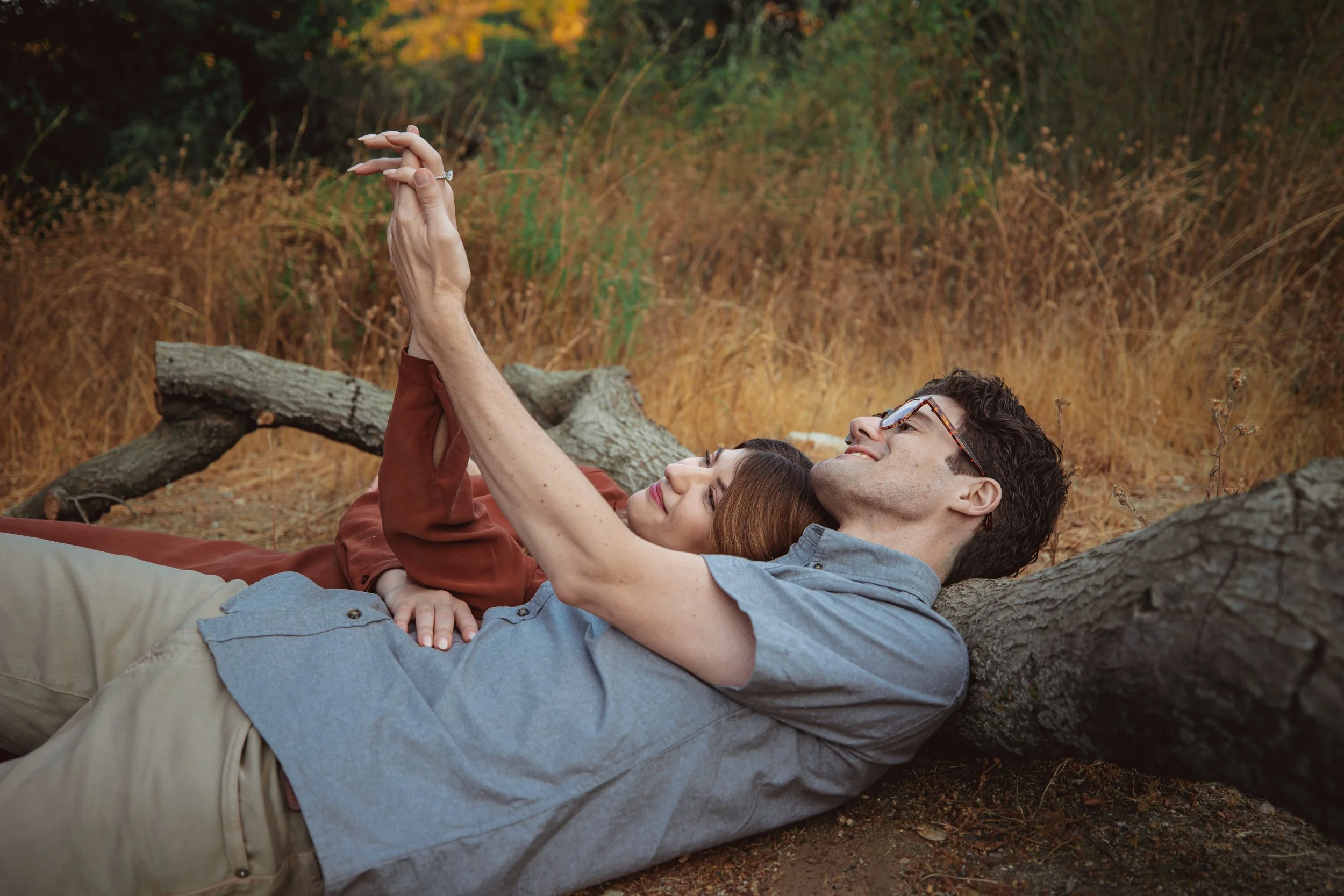 A couple lying on the ground outdoors, looking at a smartphone together, surrounded by autumn foliage and fallen leaves.