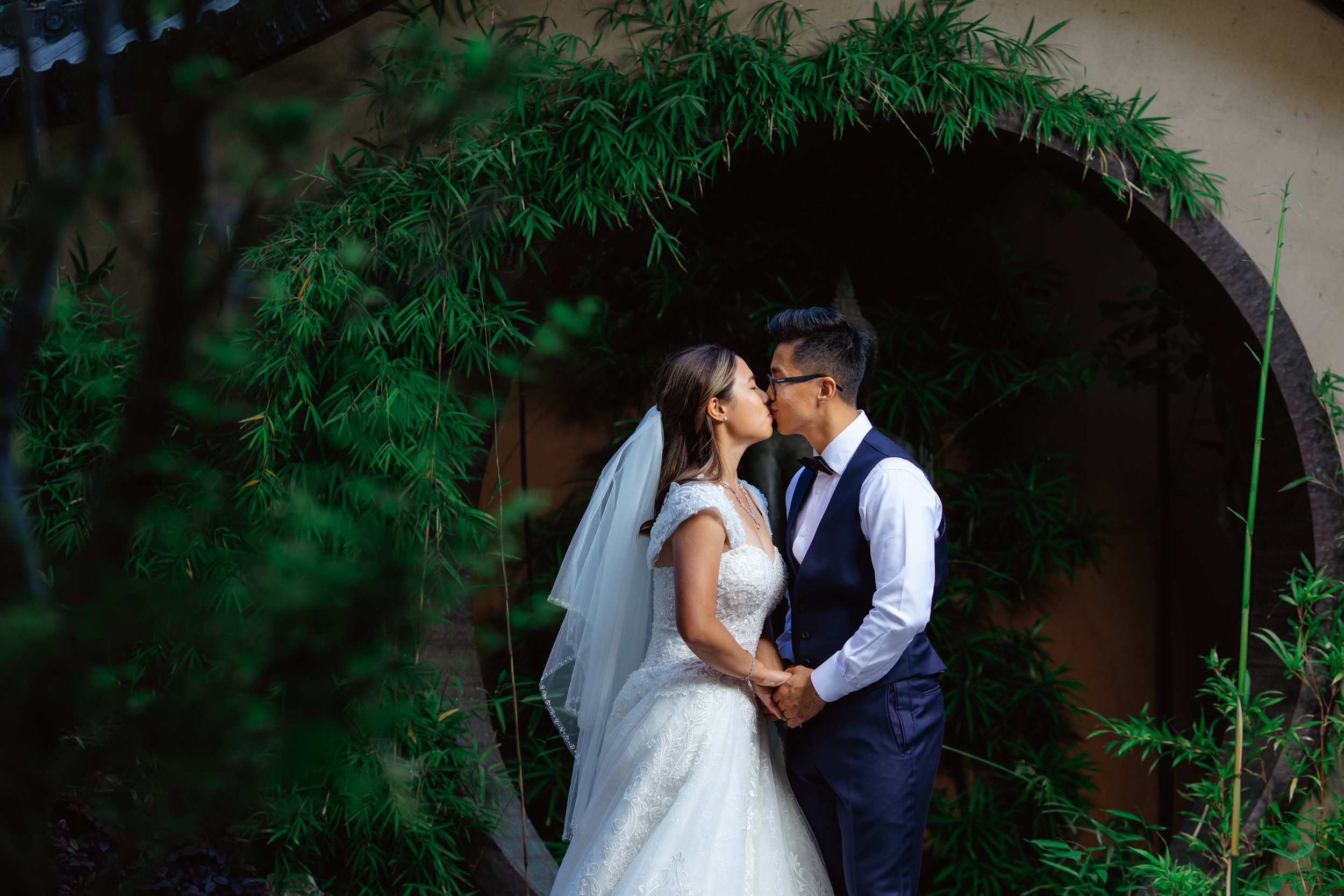 A newlywed couple sharing a kiss in front of a green leafy background with an arched opening.