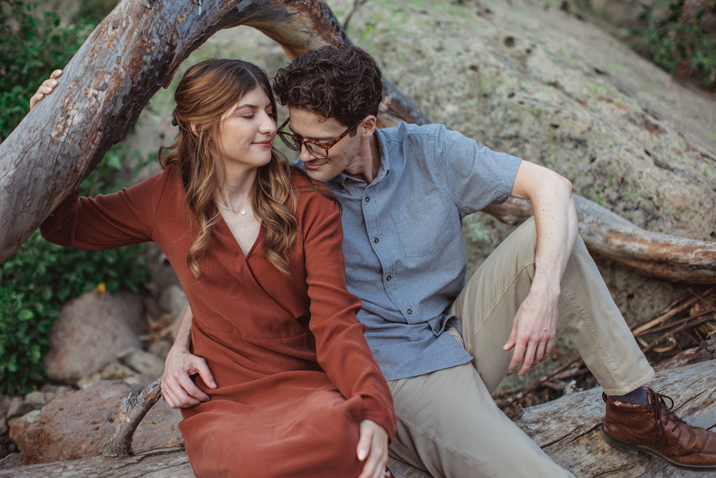 A young couple sits close together outdoors, leaning against a fallen tree with their faces touching lovingly.