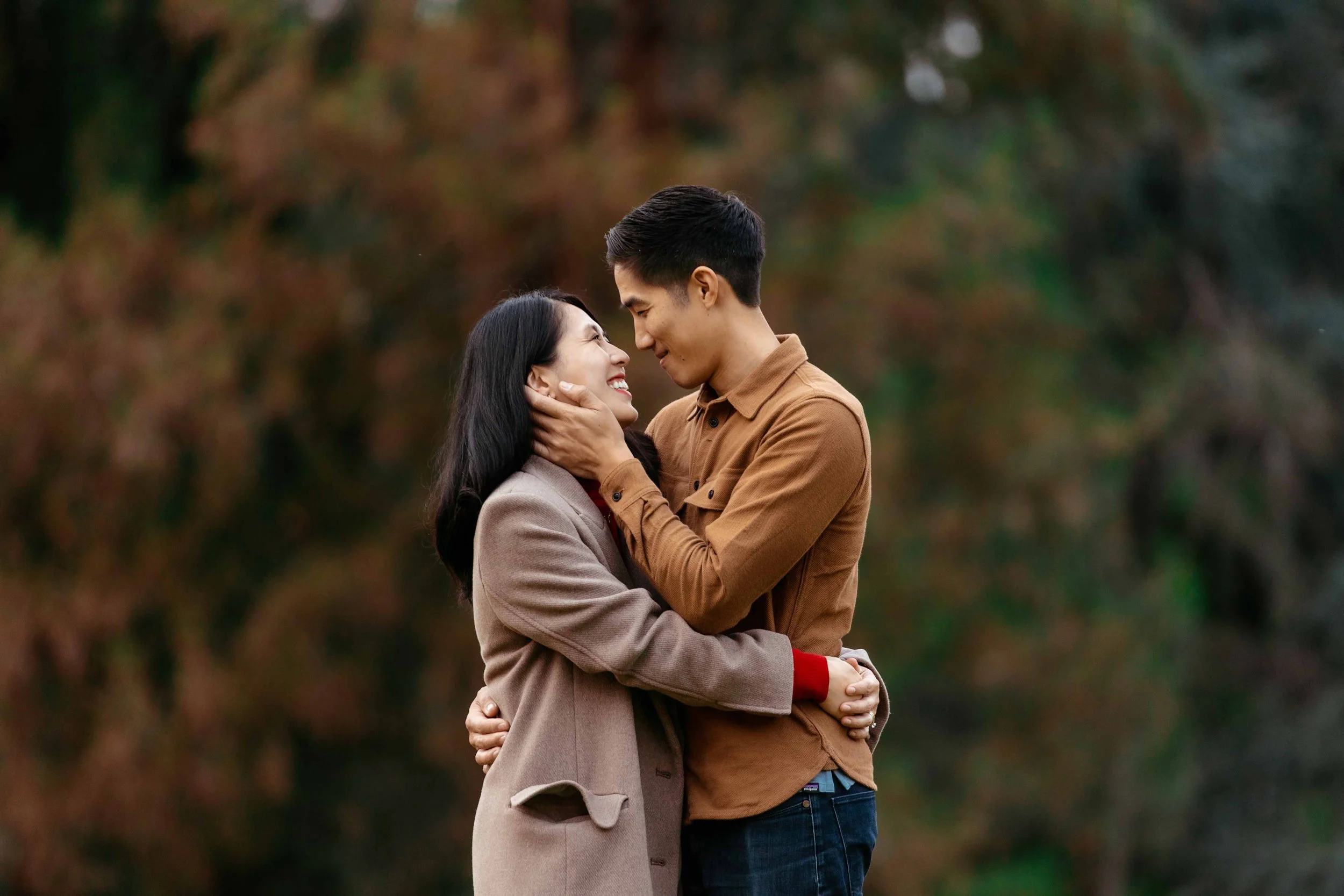 A couple smiling and embracing outdoors with blurred autumn foliage in the background.