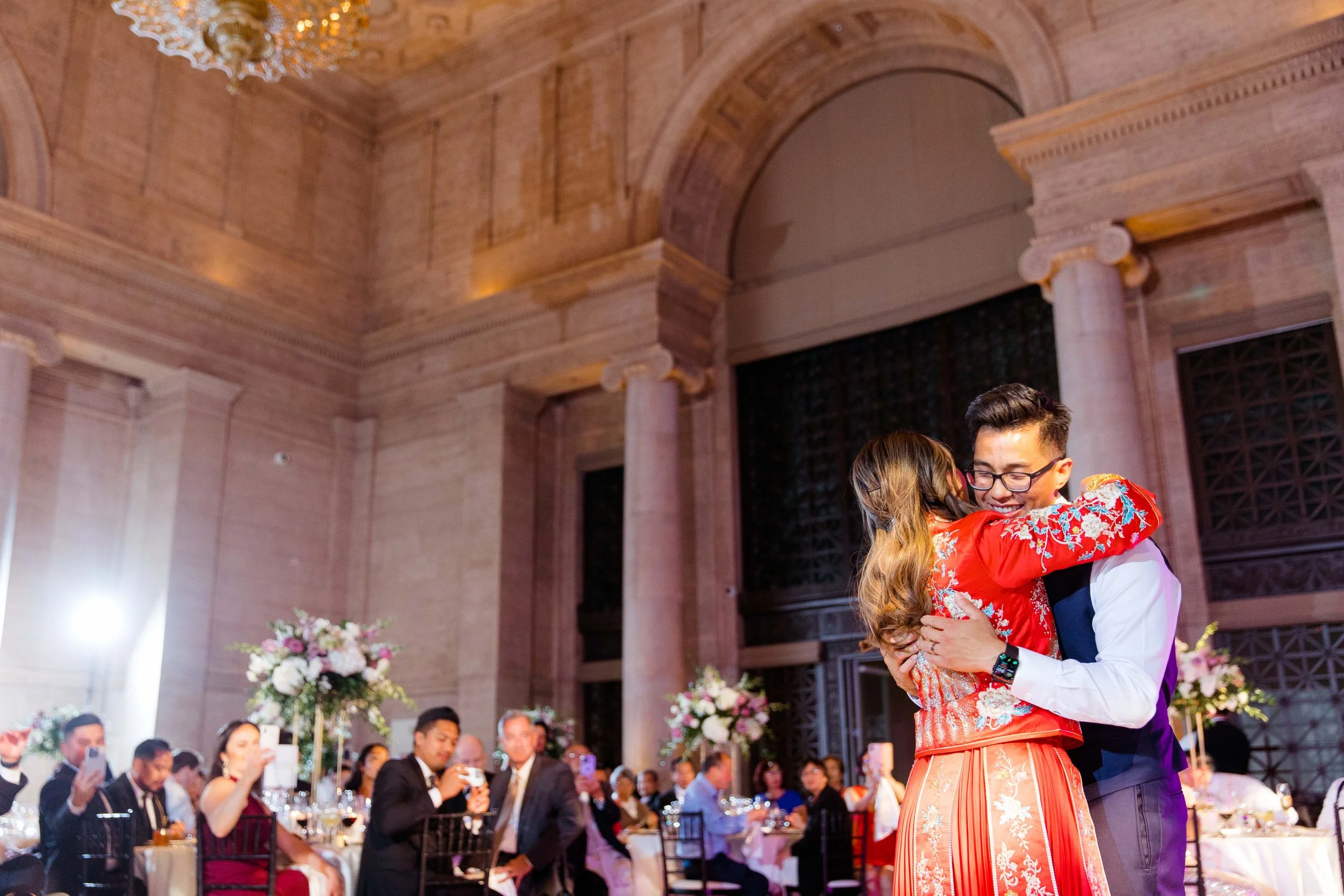 A couple hugging during a celebration in a grand, elegant hall with guests watching and taking photos.
