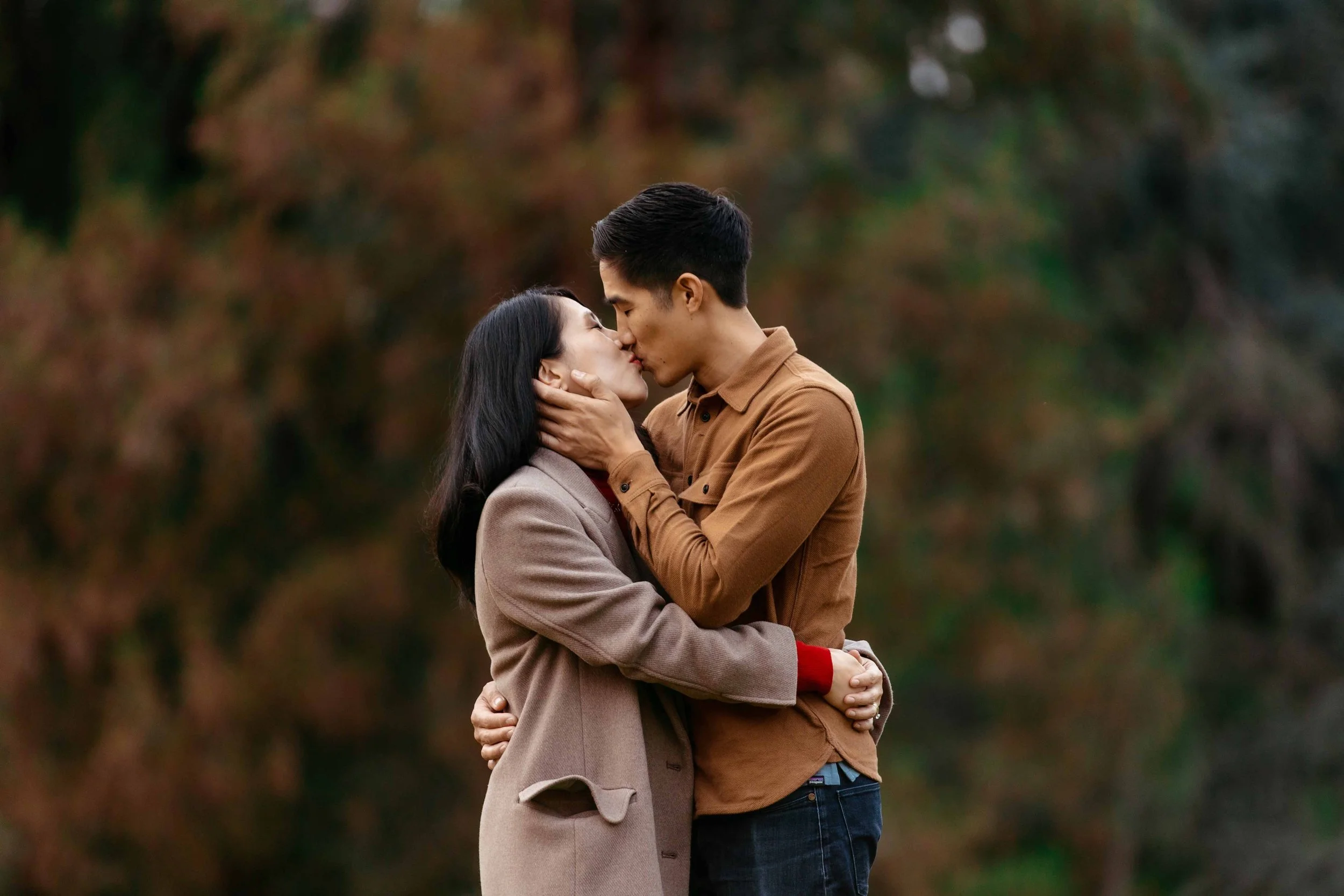 A couple sharing a kiss outdoors, embracing each other with blurred trees in the background.
