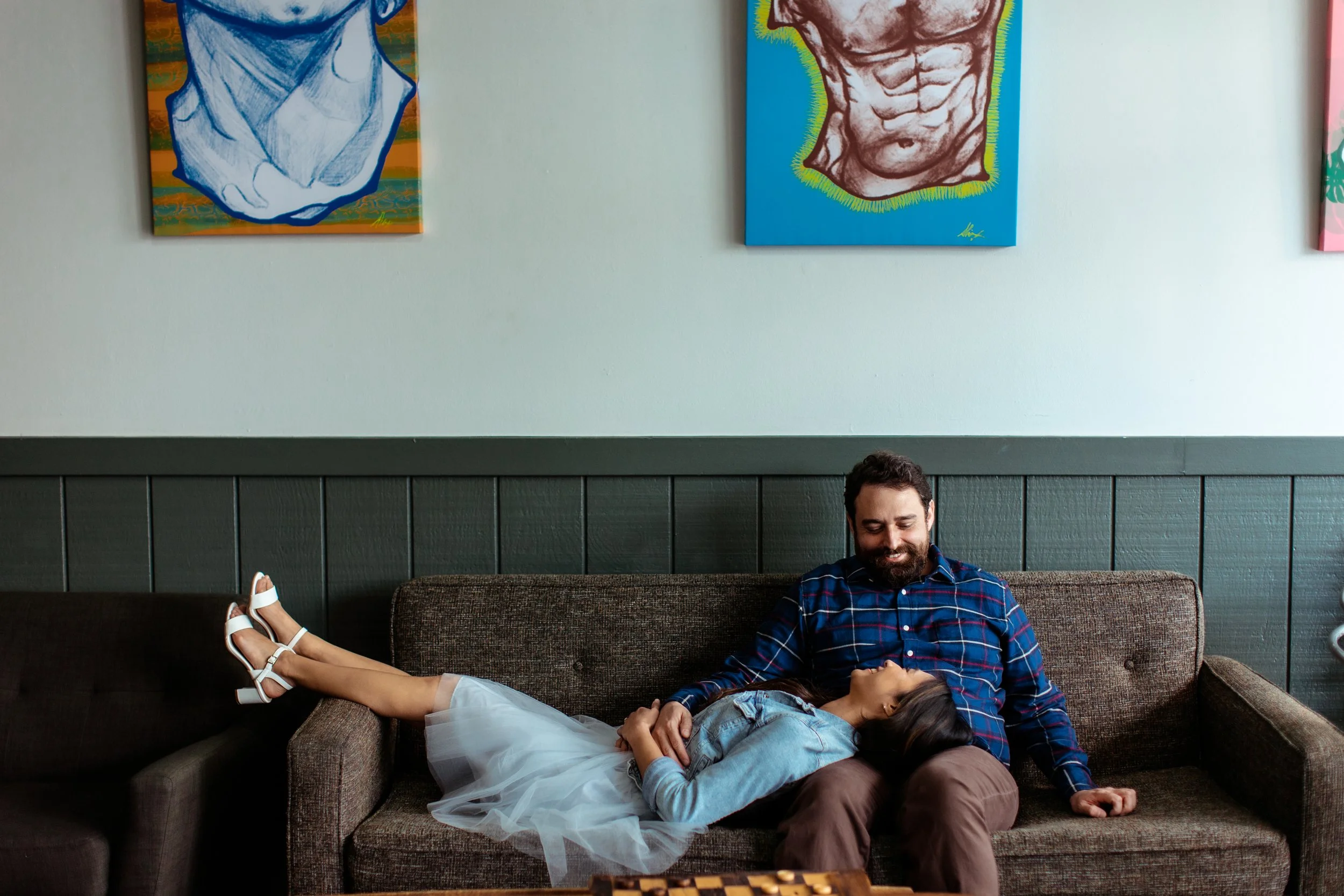 A man and a girl sitting on a brown sofa in a living room. The girl is lying across the man's lap with her feet up on an adjacent sofa. They are smiling and looking at each other. There are colorful art paintings on the wall behind them.