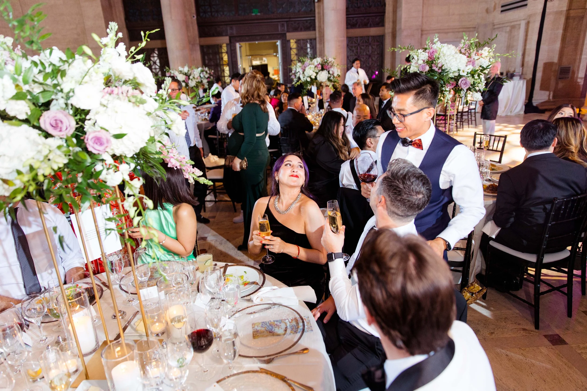 Guests celebrating at a formal event with elegant floral centerpieces, dining tables, and a server holding a glass of red wine