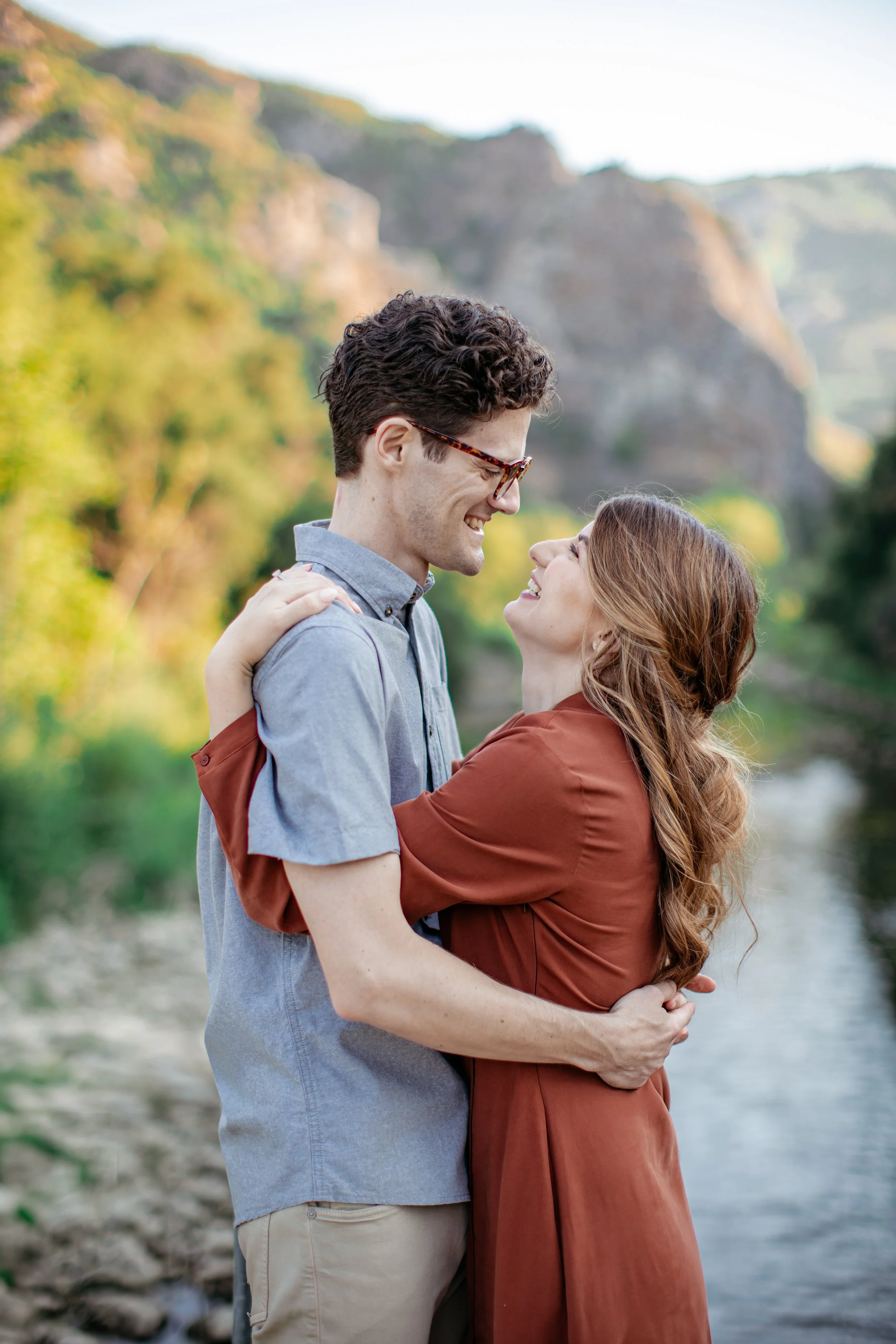 A couple embracing outdoors near a river with green trees and mountains in the background, smiling at each other.