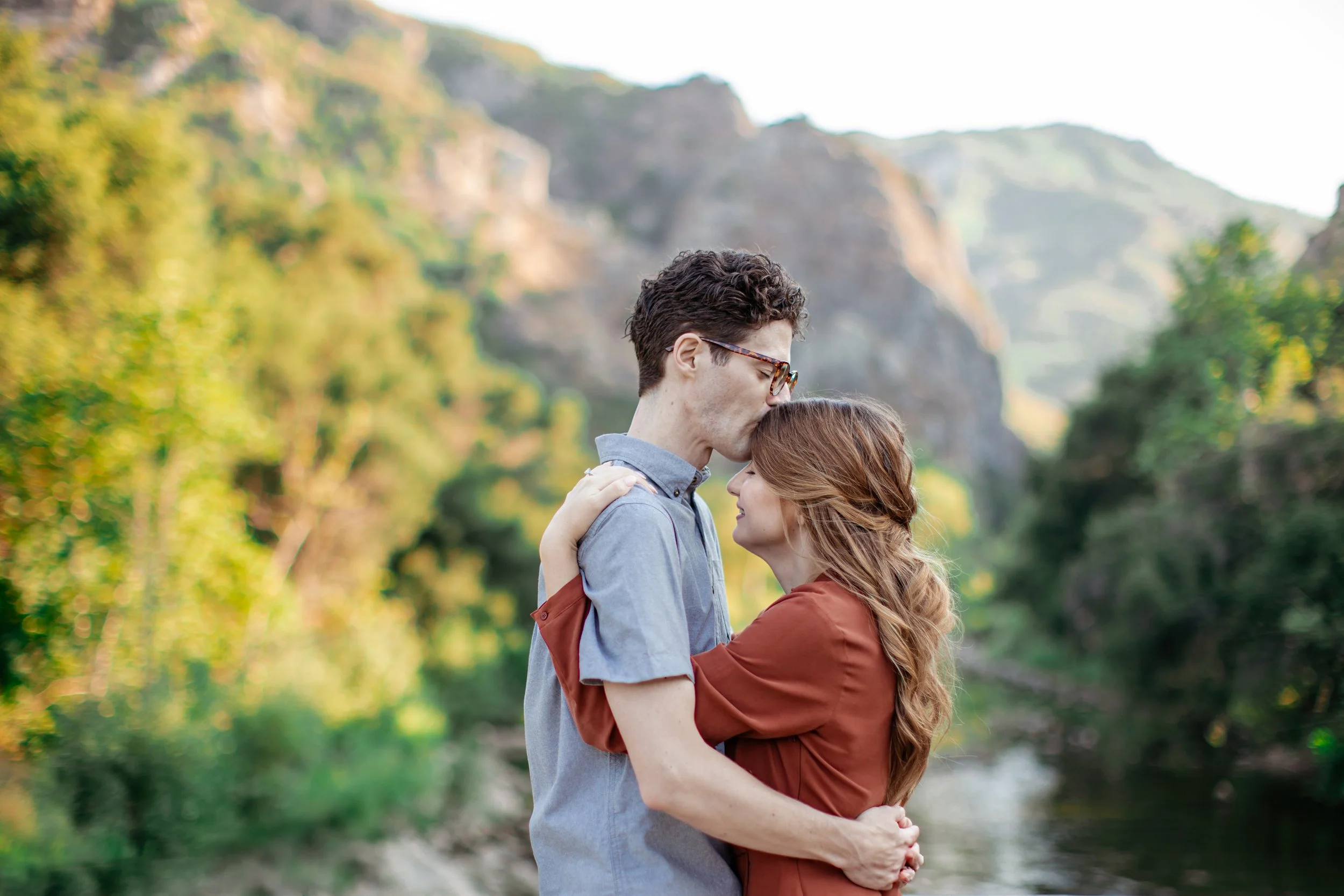 A couple embraces outdoors with the man kissing the woman's forehead, in a natural setting with greenery and rocky formations in the background.