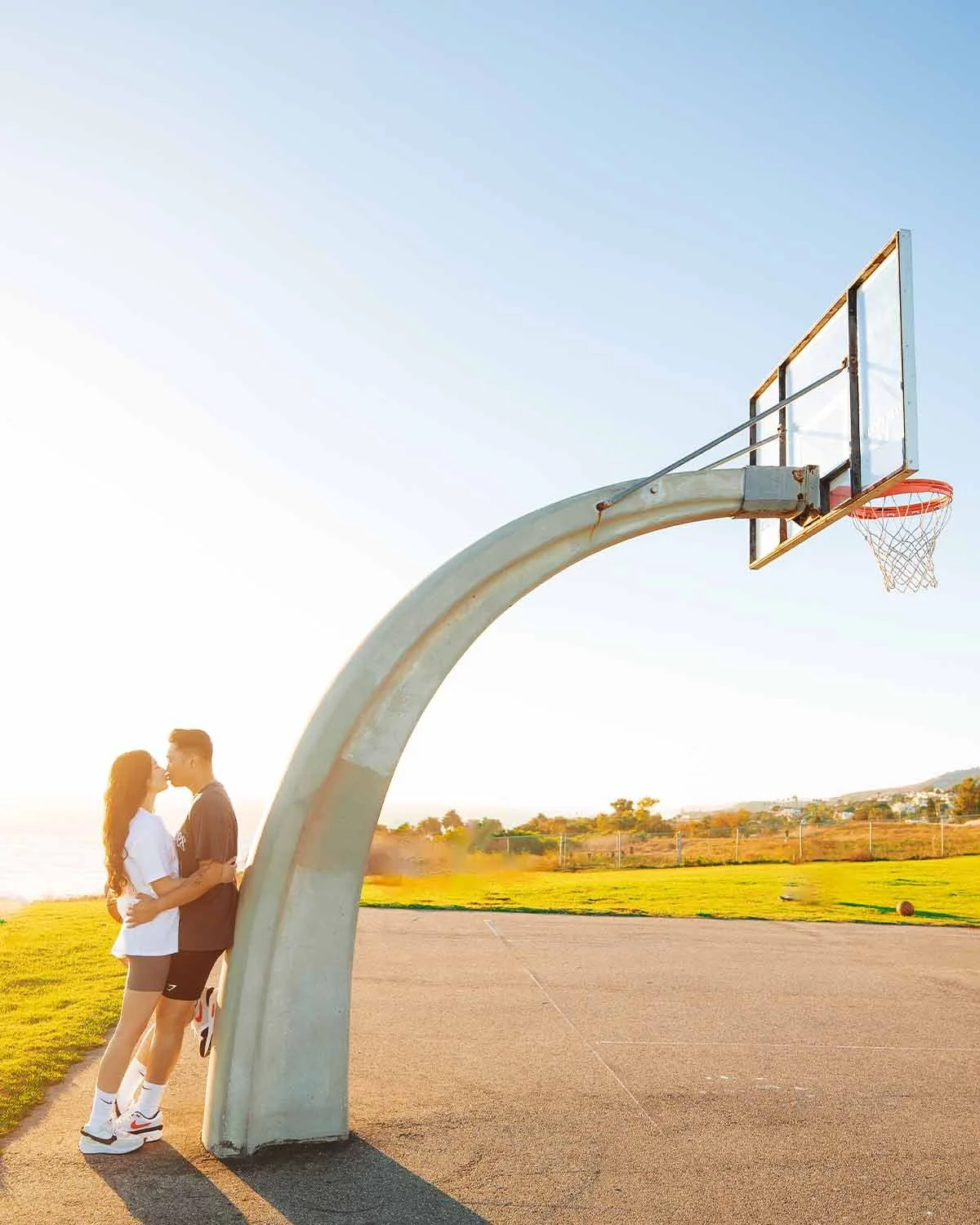 A couple stands close together on an outdoor basketball court near a basketball hoop, with the sunset in the background.