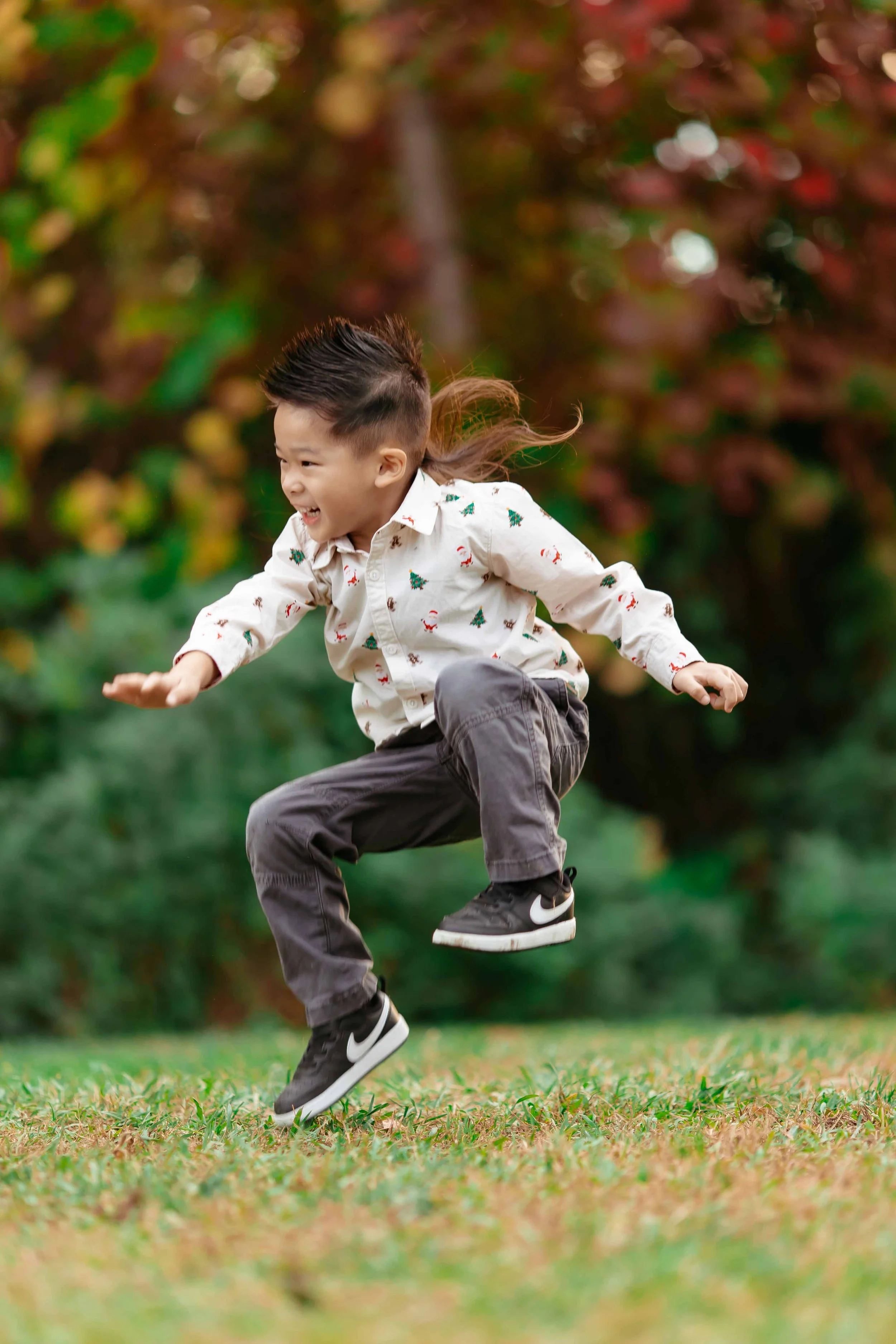 A young boy wearing a Christmas-themed shirt and dark pants jumping outdoors on grass with a background of colorful autumn trees.
