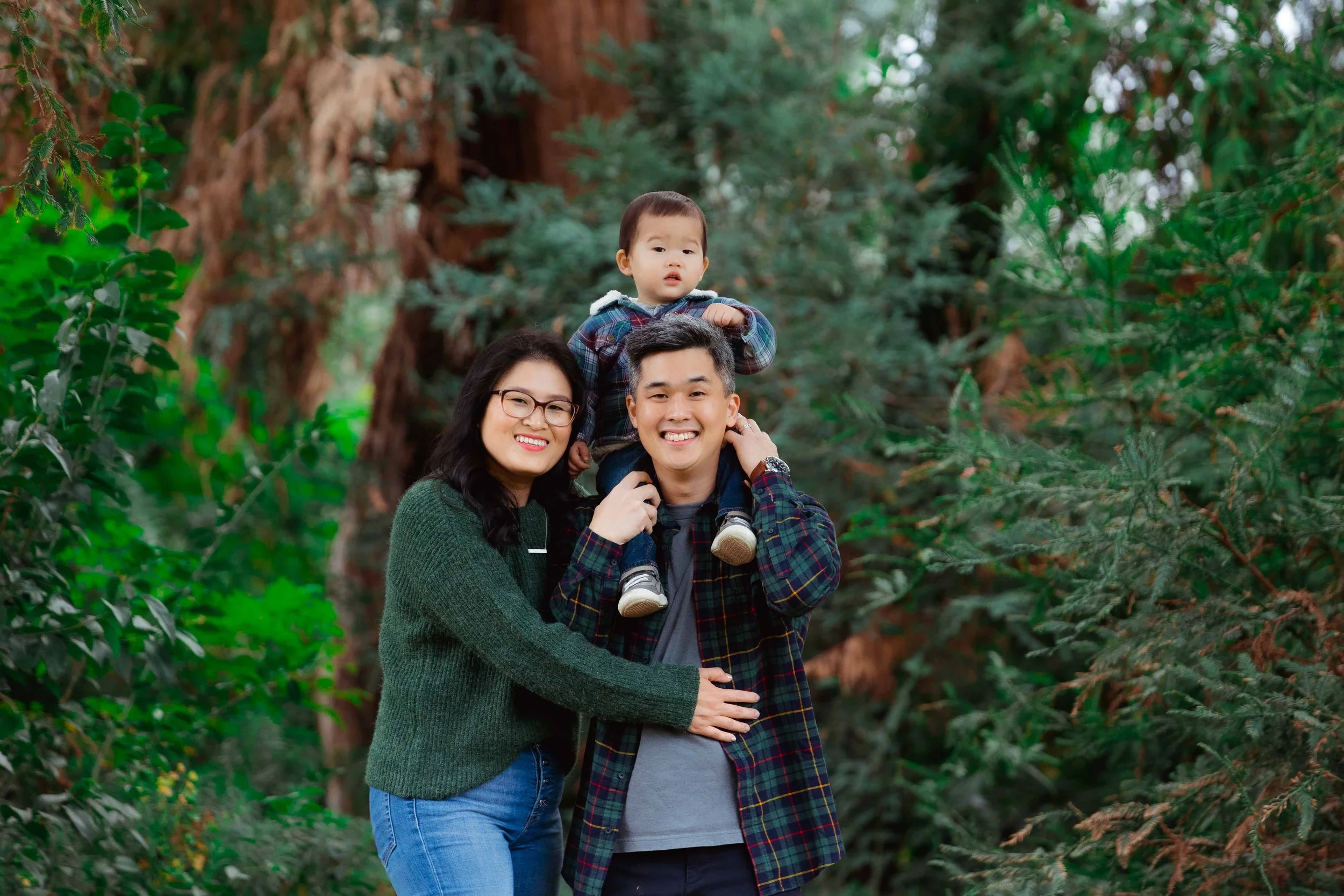 A happy family of three in a forest, with a woman wearing glasses and a green sweater, a man wearing a plaid shirt, and a child sitting on the man's shoulders.